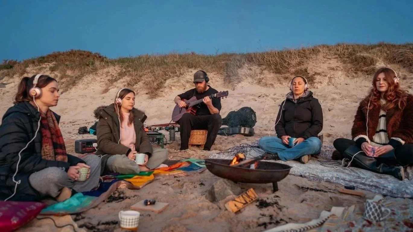 CMF participants wearing headphones around a fire during a beach session in Guernsey.