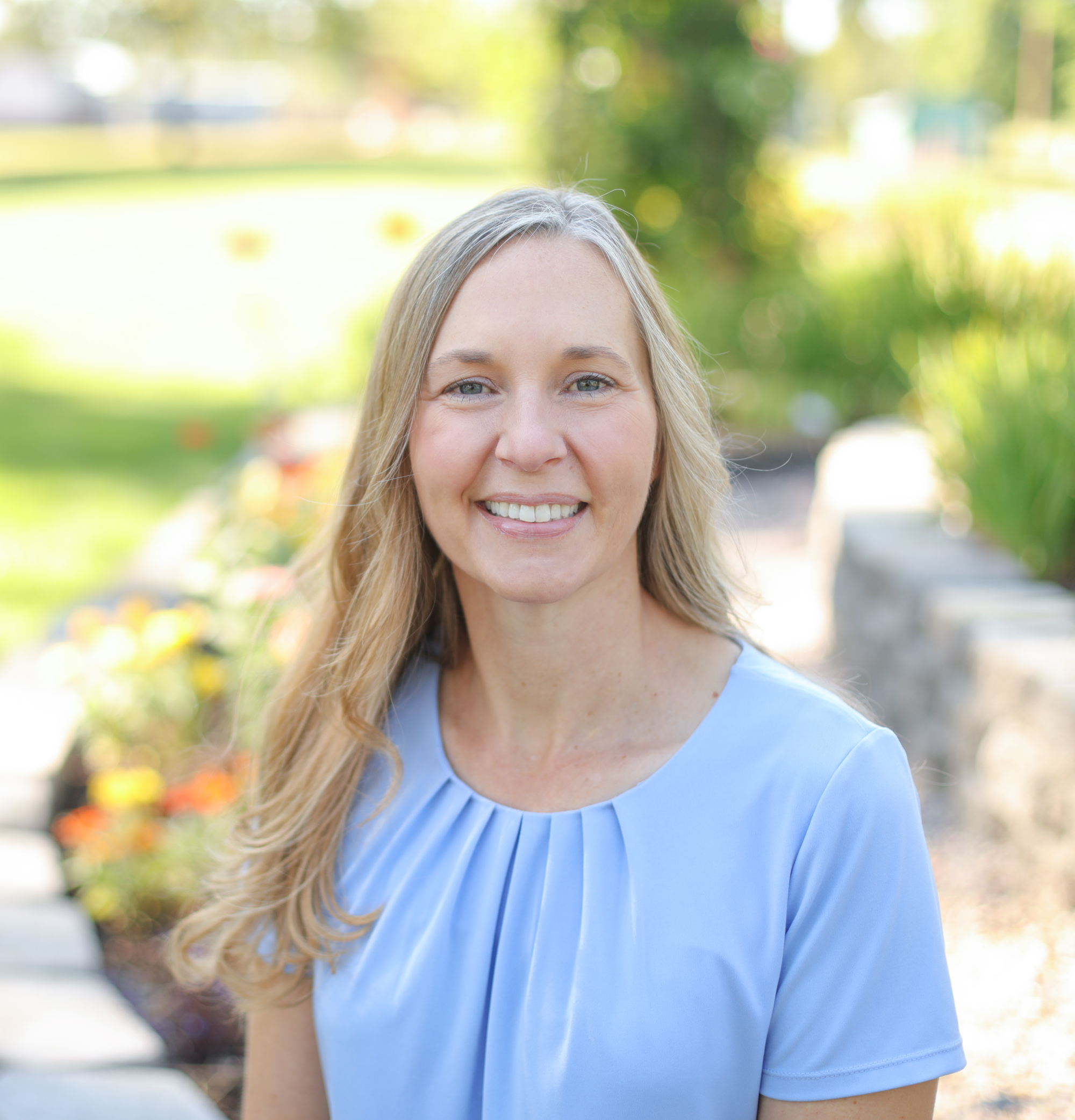 A woman with long blonde hair wearing a light blue top, smiling outdoors in a park with green trees and flowers in the background.