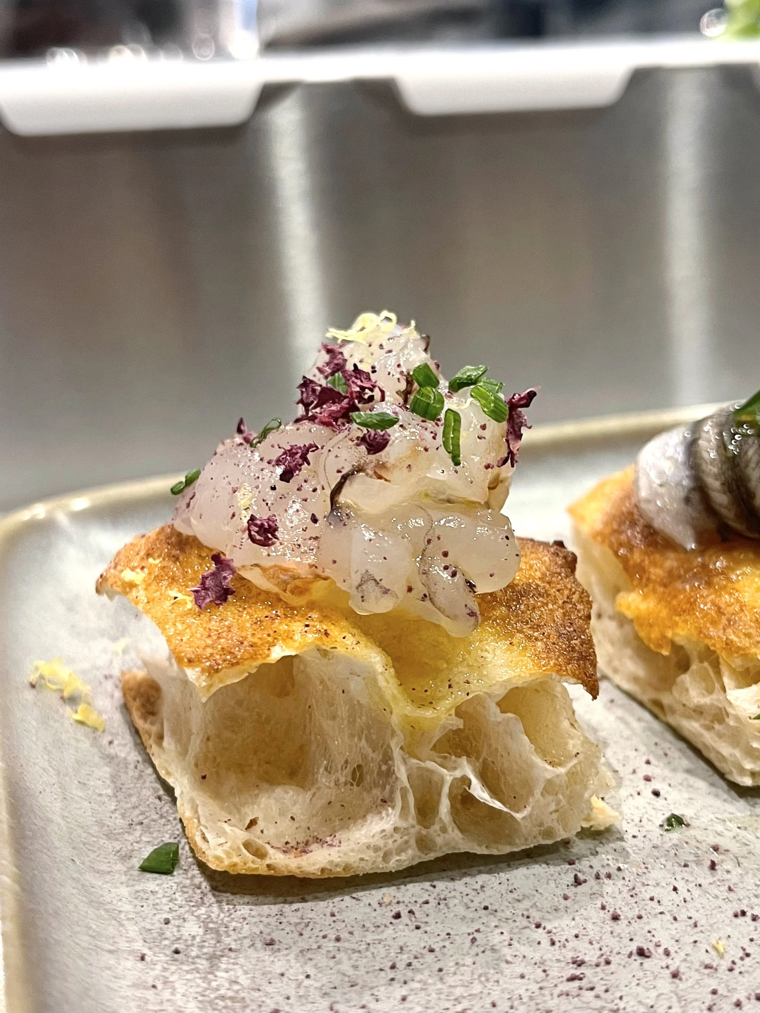 Pane tostato con copertura di pesce, erba cipollina e decorazioni di petali di fiore, in un piatto bianco.
