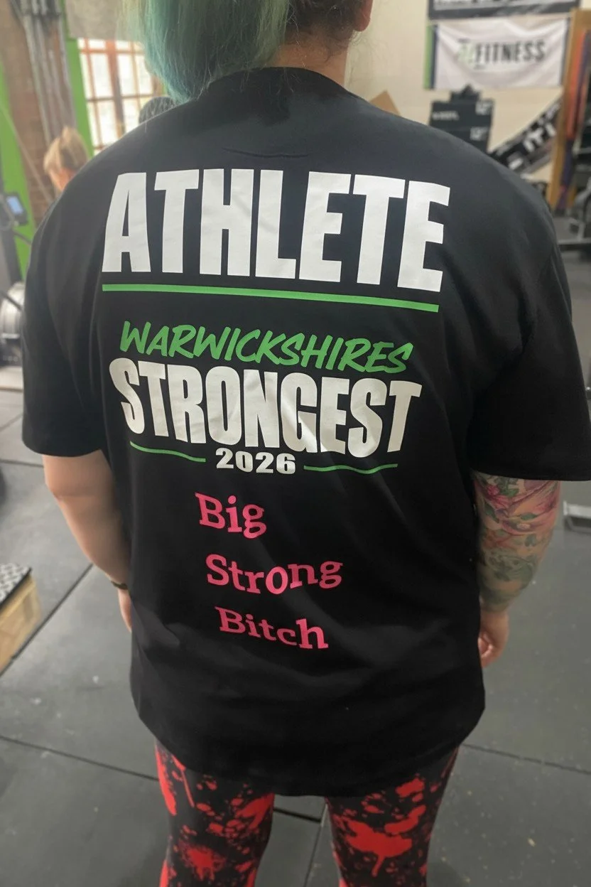 Back of a person wearing a black T-shirt with colorful text that reads "ATHLETE WARWICKSHIRES STRONGEST 2026 Big Strong Bitch." The person has a tattooed arm and multicolored hair, and is in a gym environment.