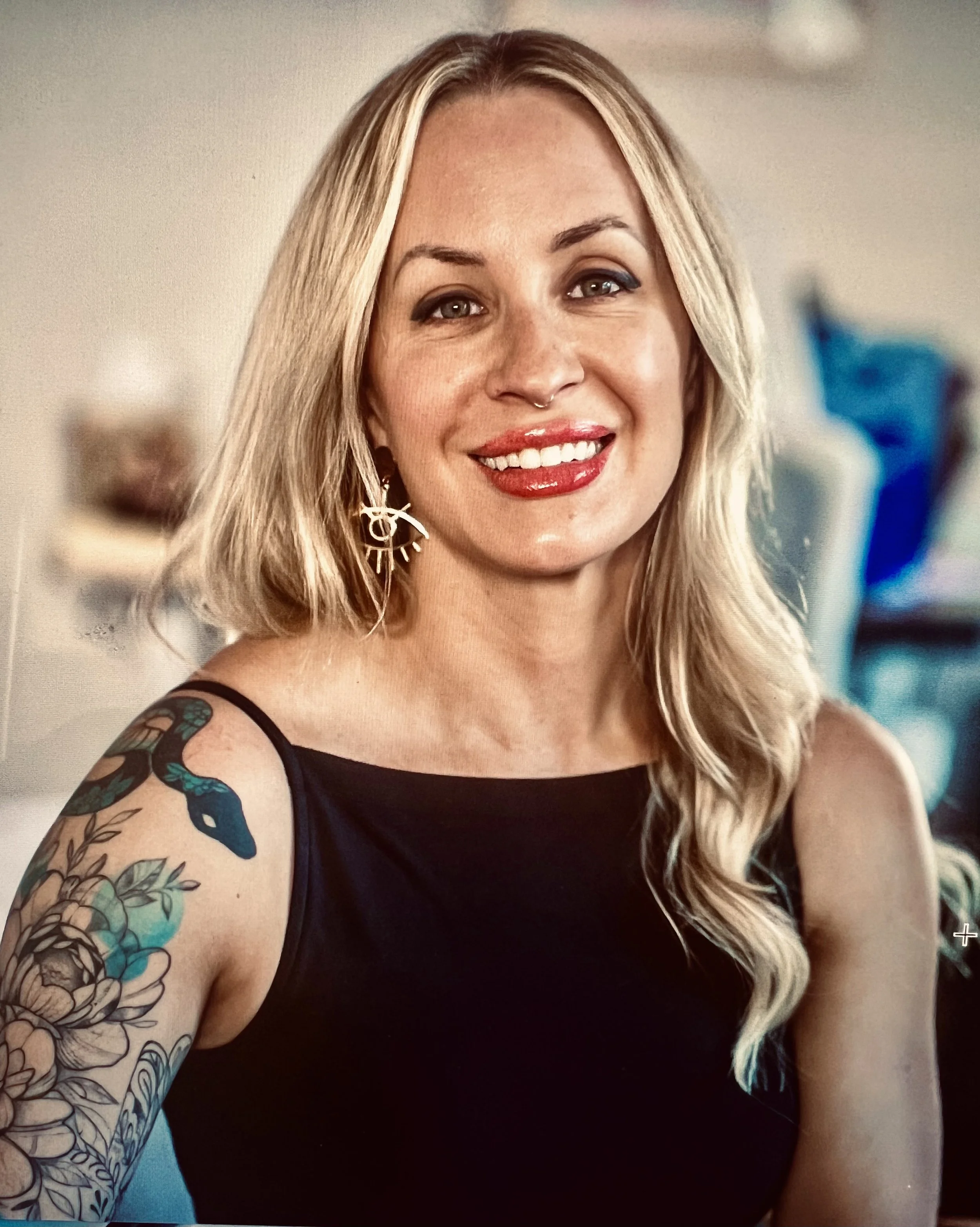 Close-up of a woman with long blonde hair, wearing a black top, and earrings. She has tattoos on her arm, red lipstick, and is smiling at the camera.