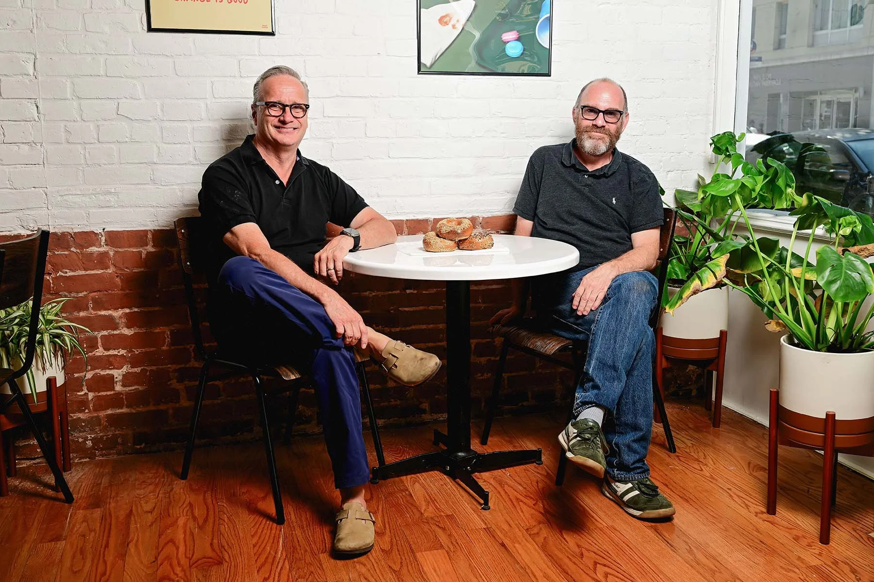 Two men sit at a small, round white table with a few bagels on it in a cafe. One man wears glasses, a black shirt, and beige slippers, while the other wears glasses, a dark gray shirt, and sneakers. There are green plants on the floor beside the window and paintings on the white brick wall behind them.