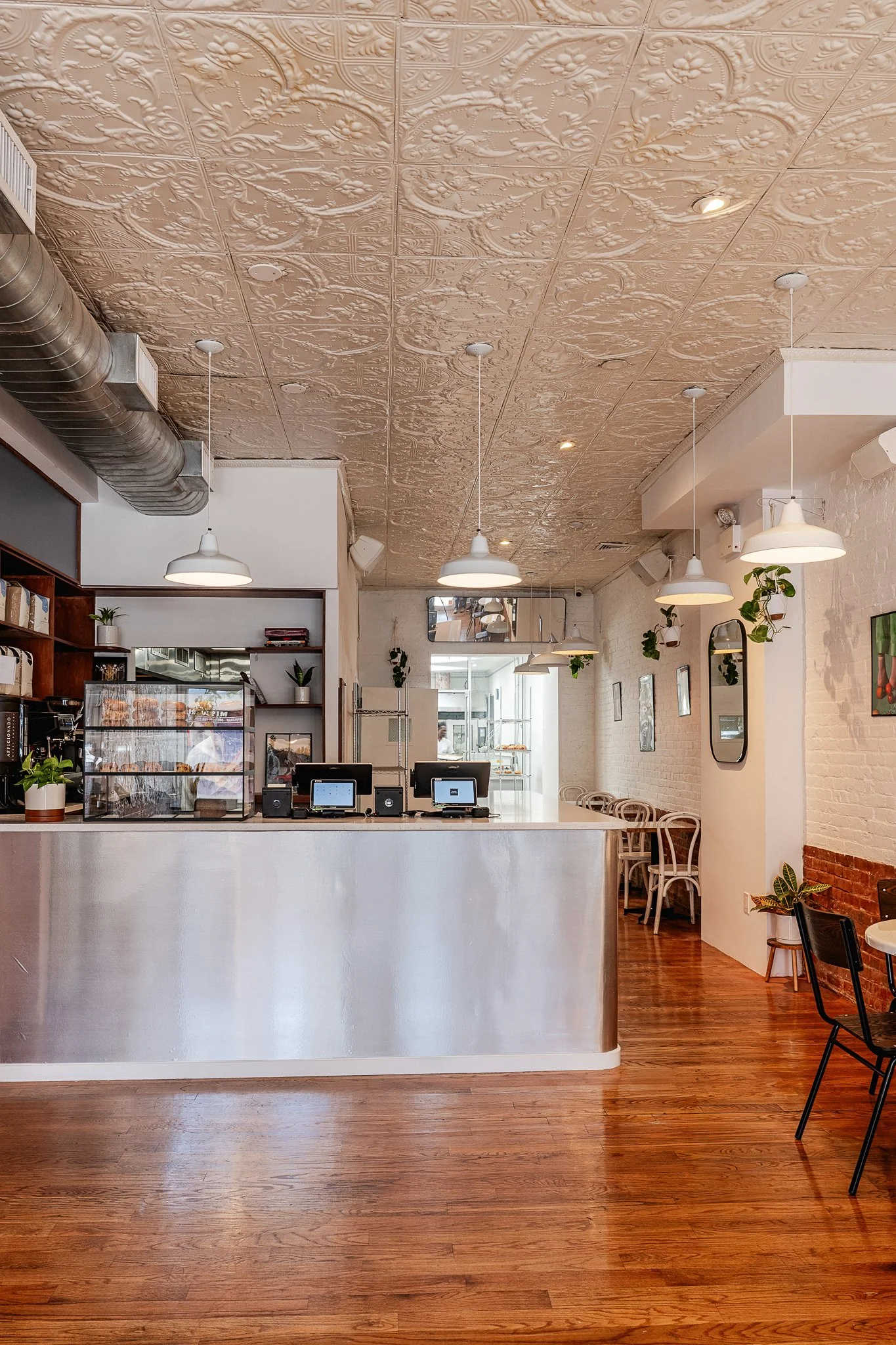 Interior of a modern cafe with a decorative ceiling featuring embossed floral patterns, hanging white pendant lights, a metal ventilation duct, and a counter with electronic payment terminals.