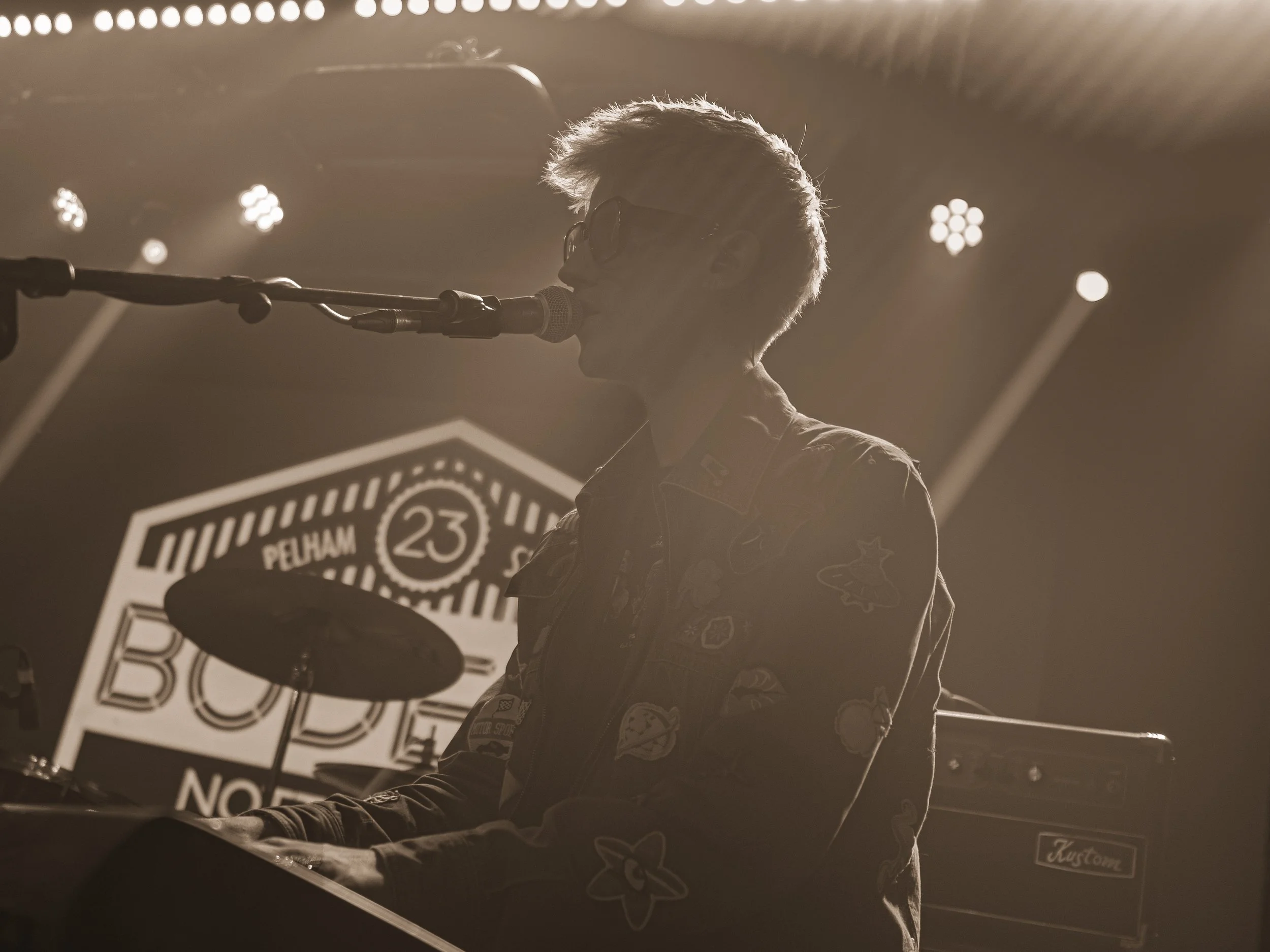 A musician wearing glasses and a patterned jacket playing a guitar and singing into a microphone on stage, backlit by stage lights.