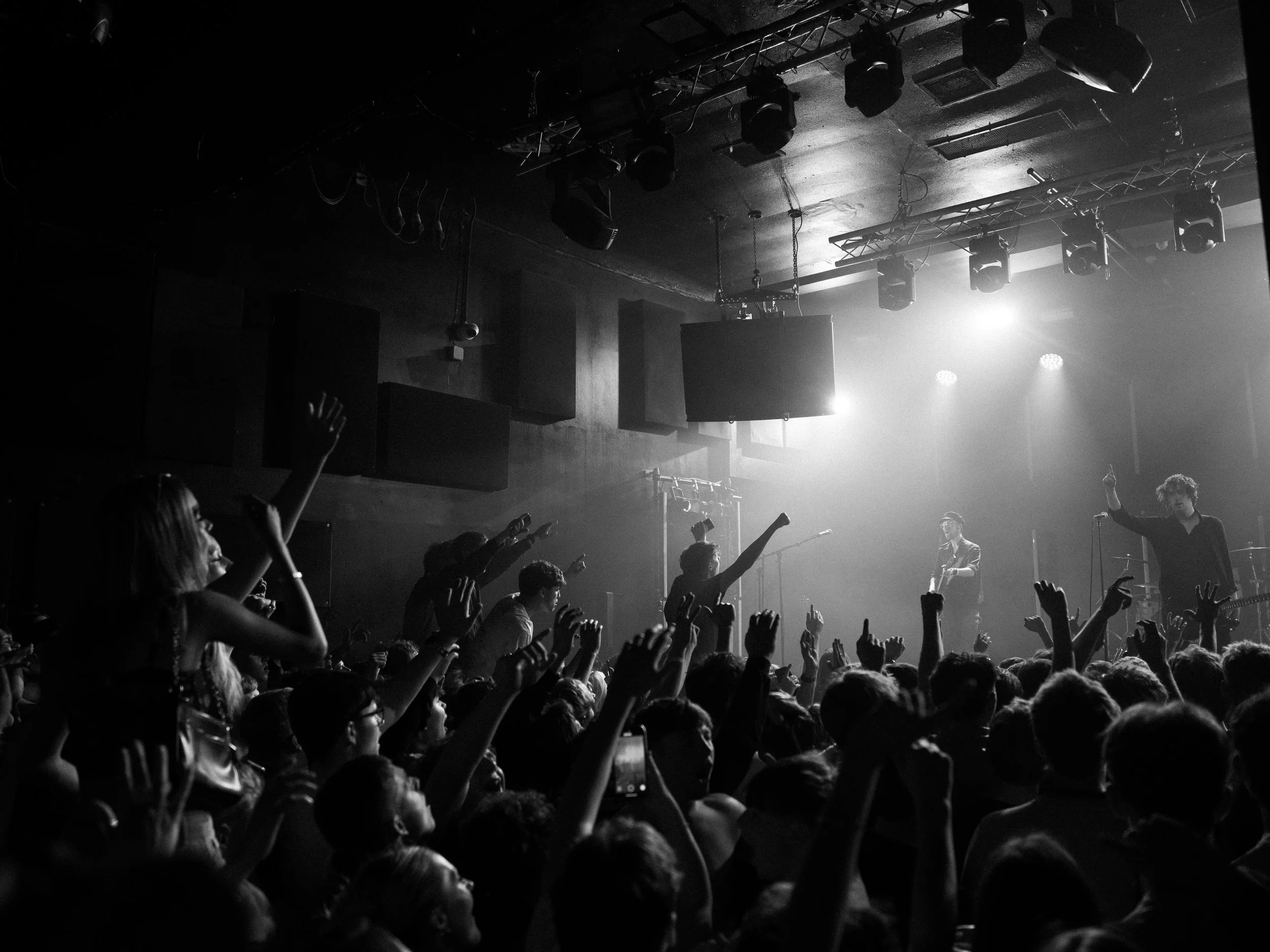 A concert or live performance with an audience raising their hands in front of a stage with performers, stage lighting, and equipment in a dimly lit venue.