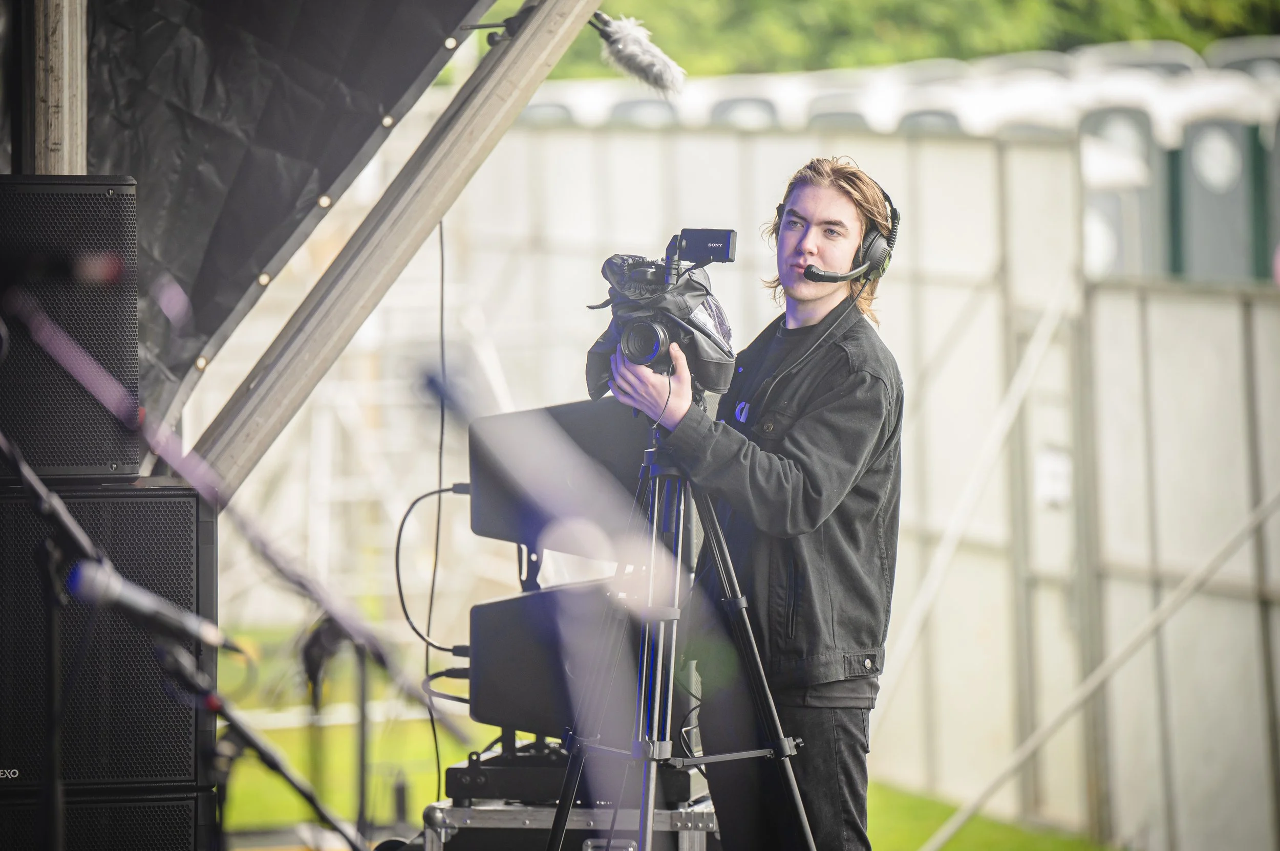 A young man wearing a black jacket and headphones operates a video camera on a tripod at an outdoor event, with a microphone attached to his head and electronic equipment nearby.