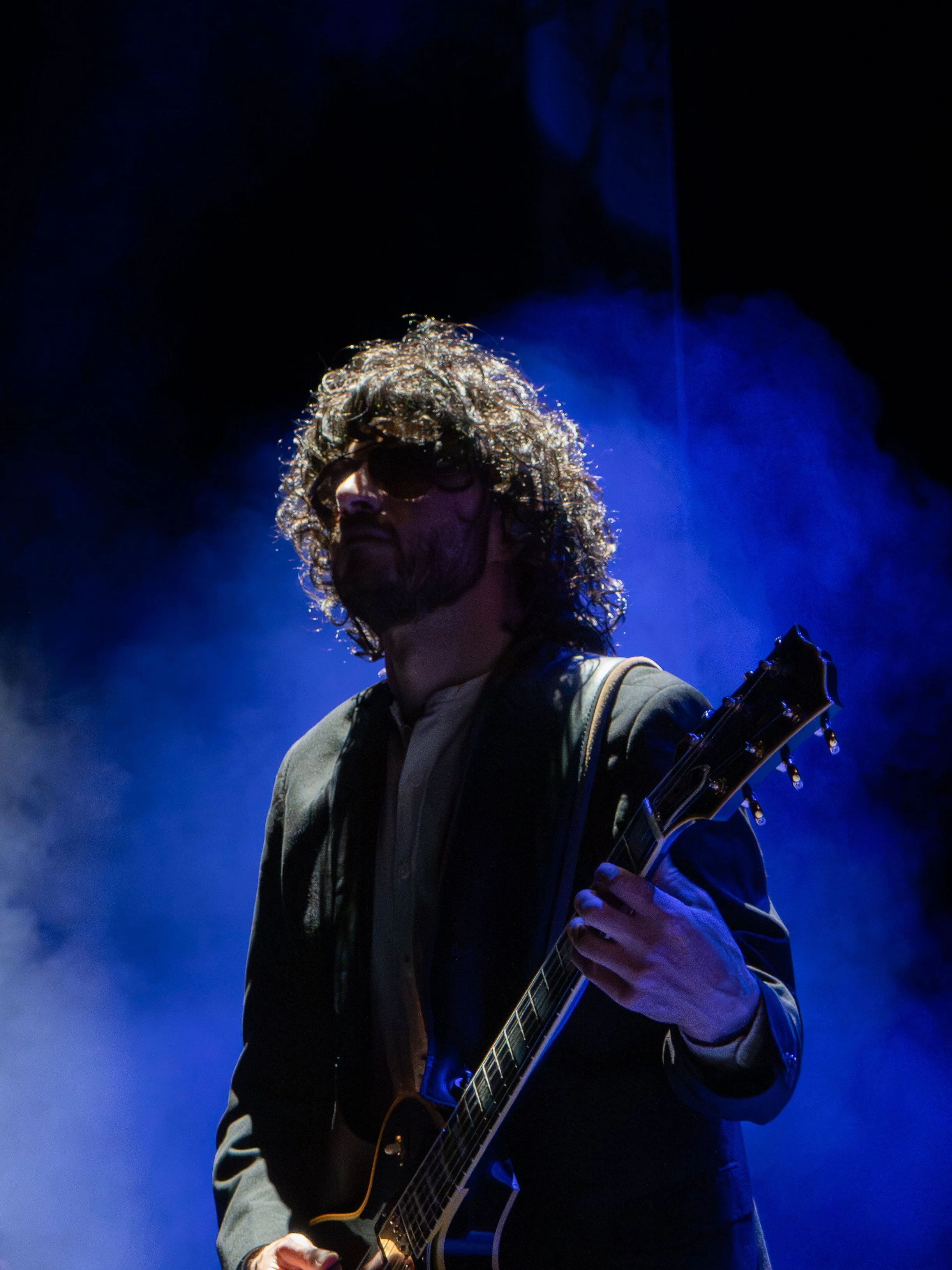 A person with curly, wet hair wearing sunglasses, playing an acoustic guitar on stage with blue lighting and smoke in the background.