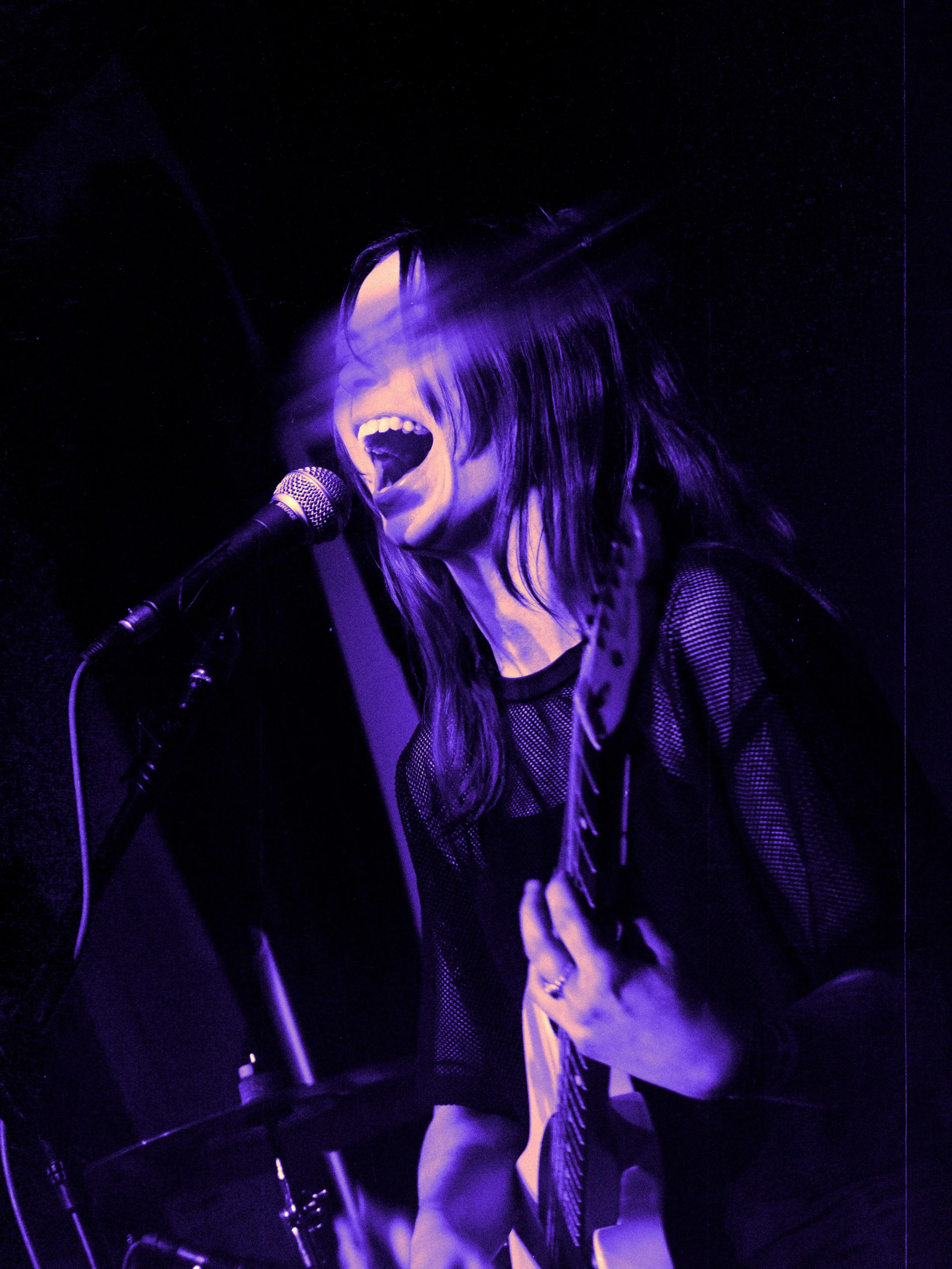 A woman singing passionately into a microphone with closed eyes and an open mouth, illuminated with purple and blue lighting, suggesting a live music performance.