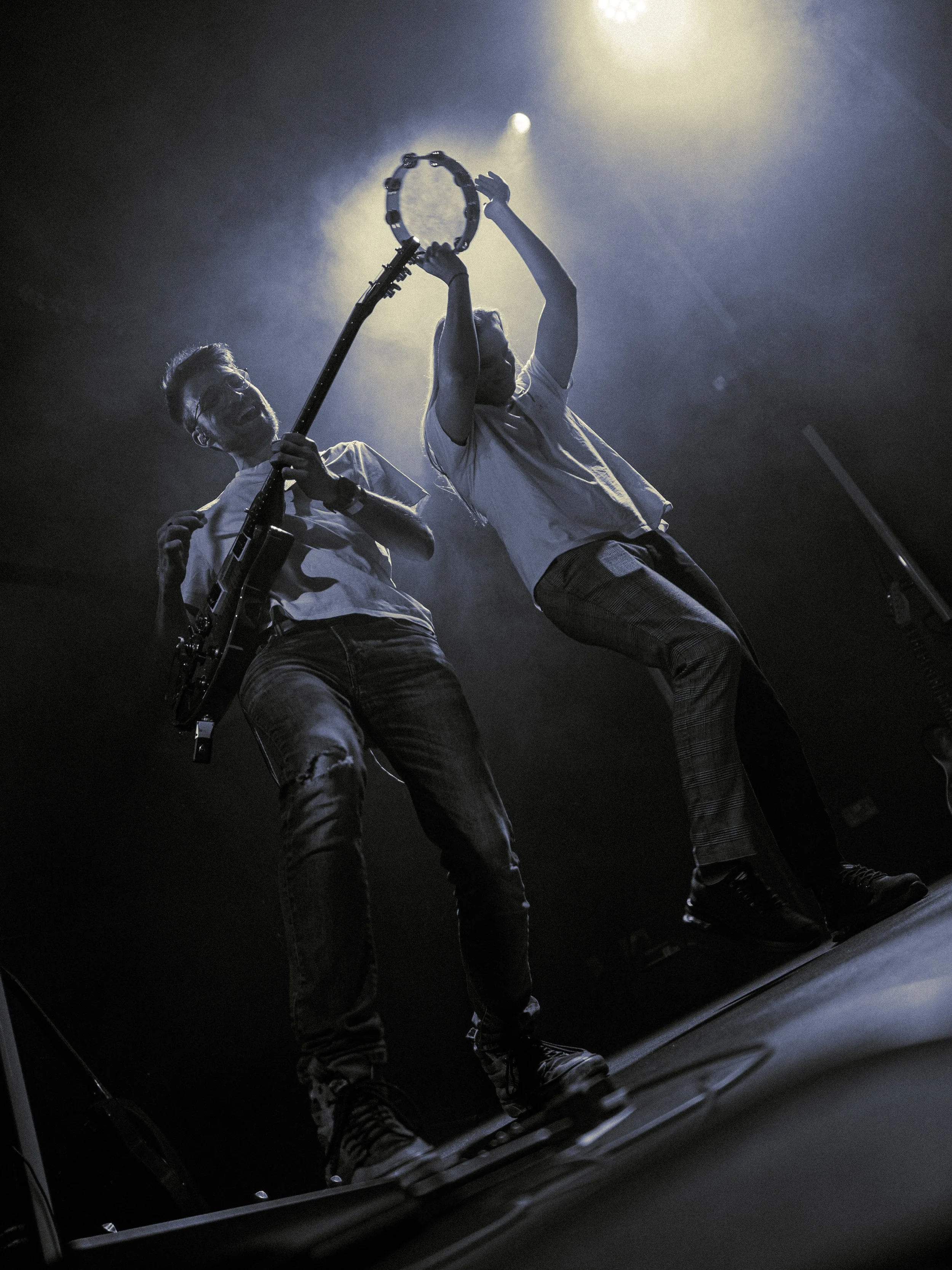 Two musicians, one playing an electric guitar and the other playing a tambourine, perform on stage under bright lights, with their reflections visible in a mirror.