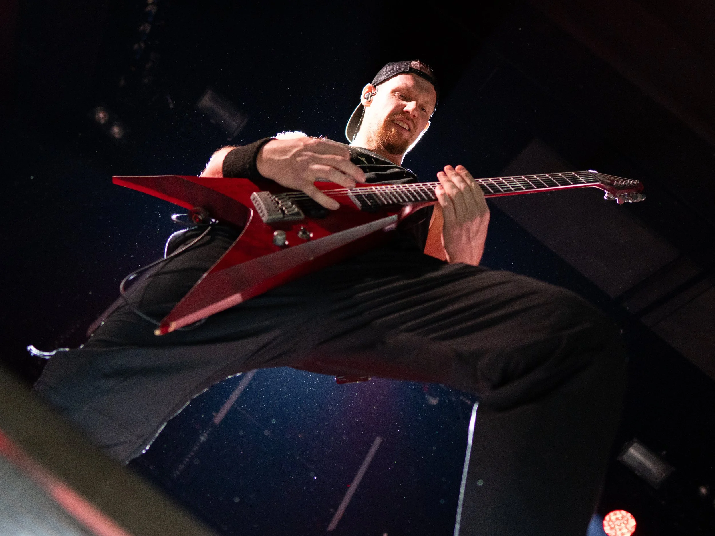 A man with a beard and a black cap, wearing a black wristband, is playing an electric guitar on stage, illuminated by stage lights against a dark background.