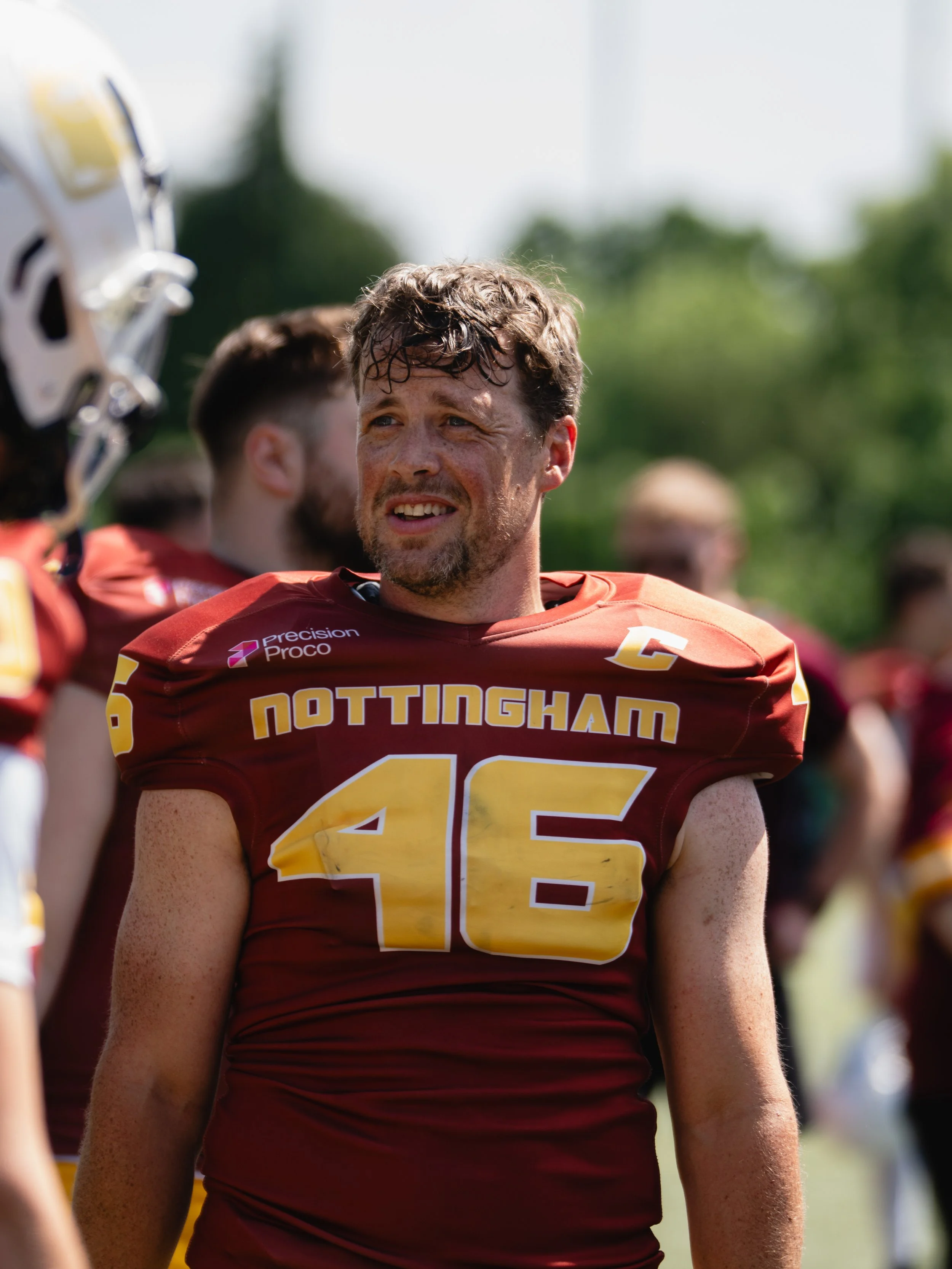 A football player with a beard and curly hair, wearing a maroon and gold jersey with 'NOTTINGHAM' and the number 15, standing outdoors with teammates in the background.