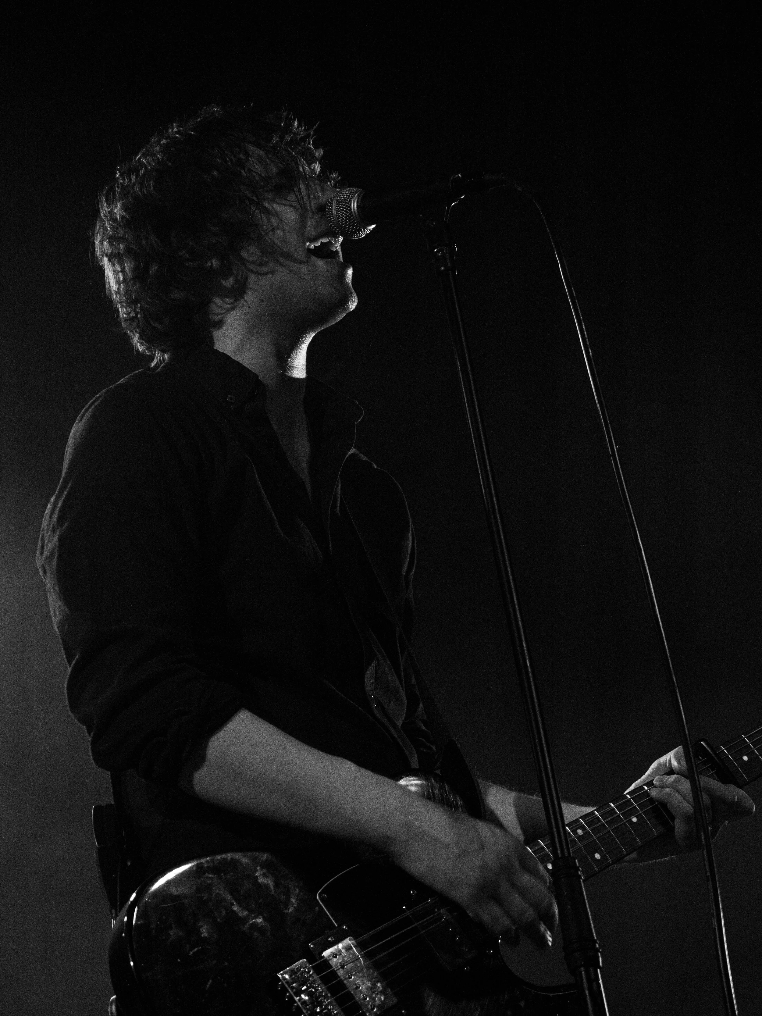 A black and white photo of a musician passionately singing into a microphone while playing an acoustic guitar. The musician has curly hair and is wearing a dark shirt. The background is dark and the focus is on the performer.