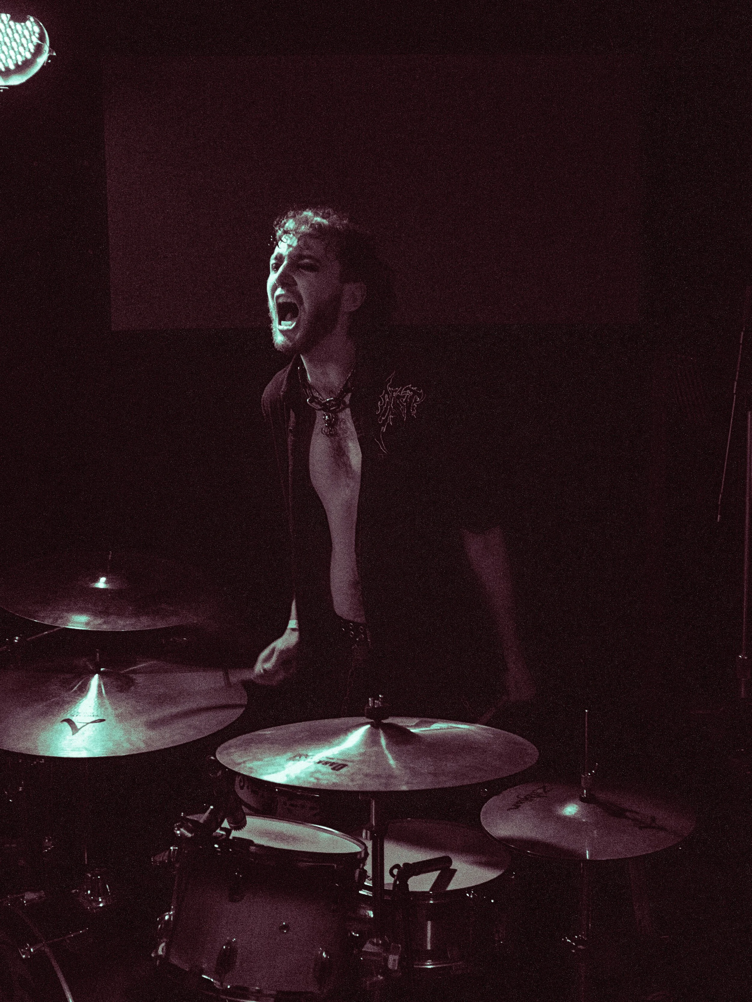 A man with curly hair and glasses screaming or singing passionately, wearing a black shirt with embroidery, behind a drum set in a dimly lit setting.