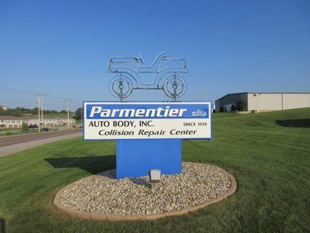 Business sign for Parmentier Auto Body Inc., collision repair center, with a metal outline of a vintage car on top, located on a grassy area near a road and industrial building.