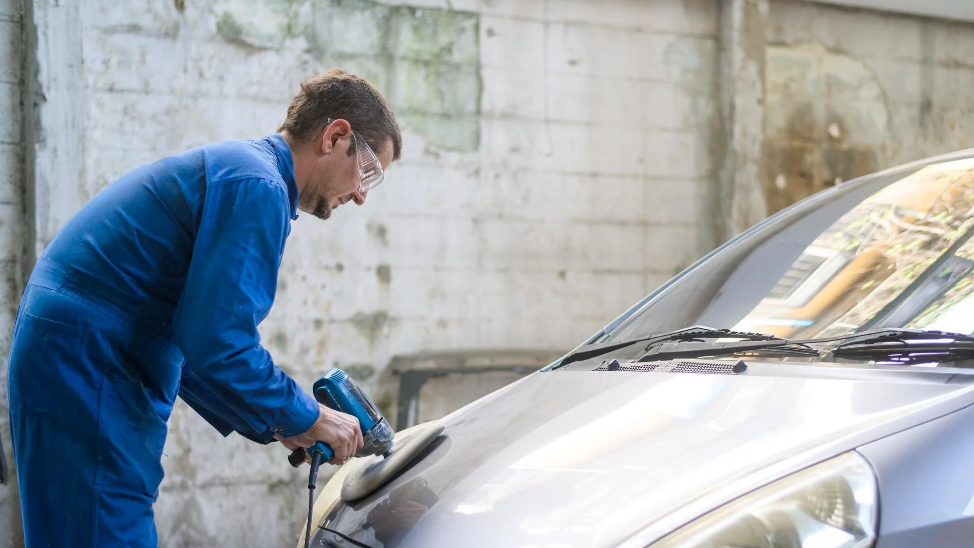 A mechanic wearing safety glasses and a blue coverall working on a car engine in a garage.