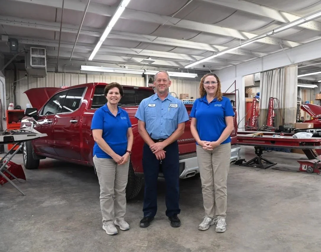 Three people standing in a garage workshop in front of a red pickup truck with its hood open, surrounded by various tools and equipment.