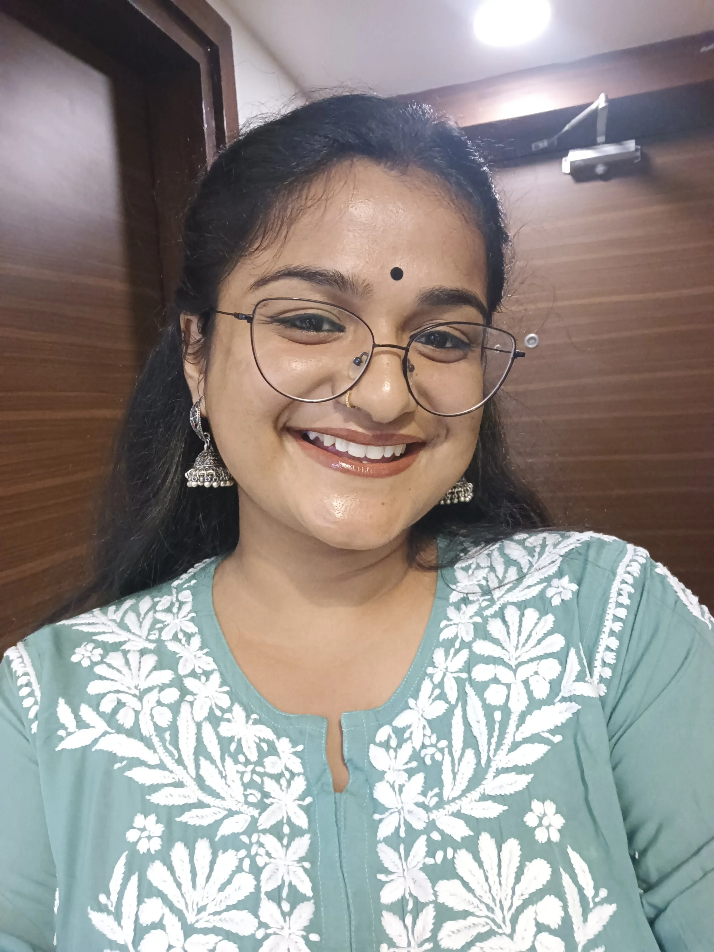 A woman smiling with glasses and traditional earrings, wearing a green embroidered dress, standing in front of a wooden wall and a ceiling light.