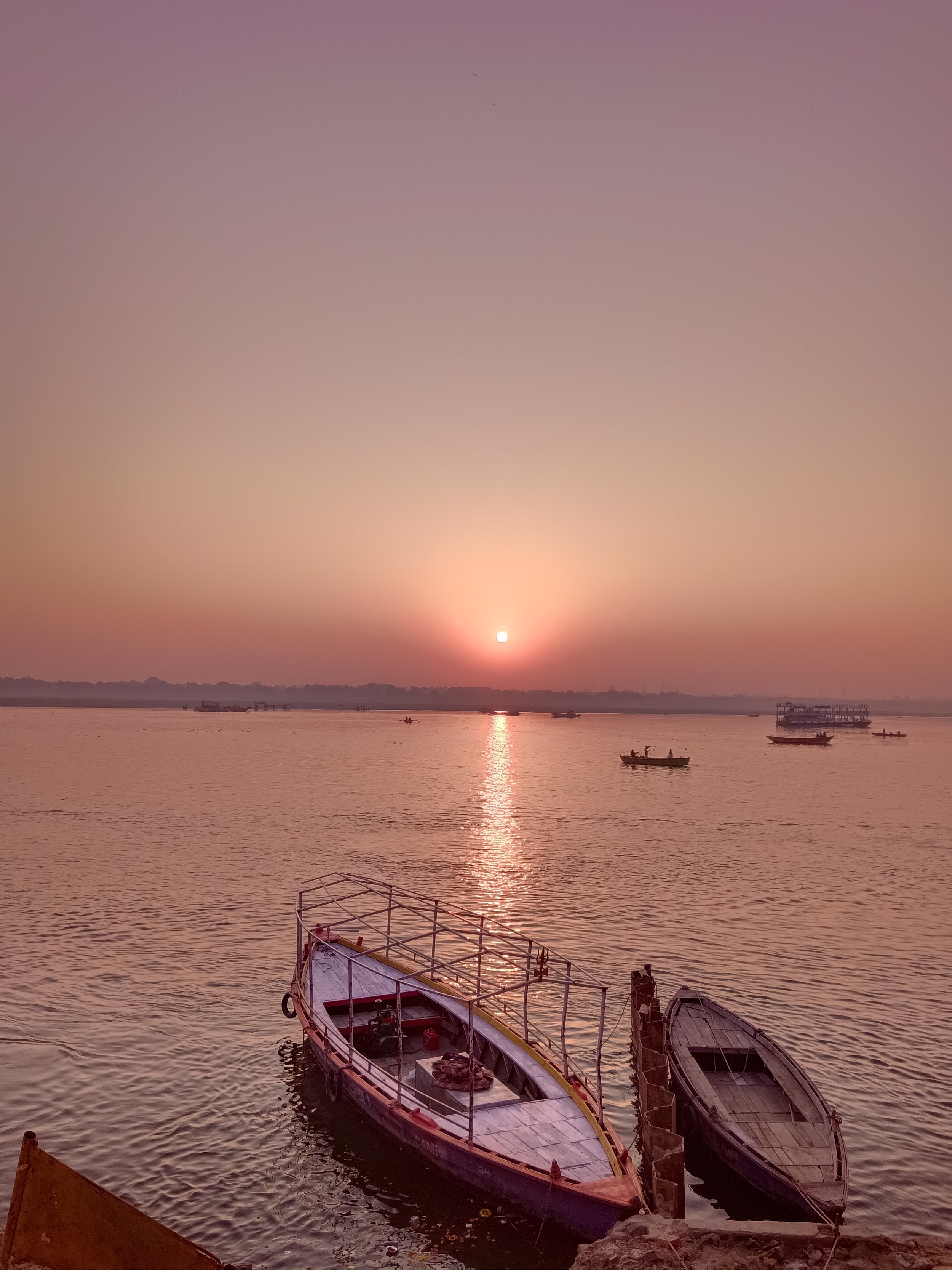 A peaceful river scene at sunset with several boats floating on the water. The sky is pinkish-purple, and the sun is near the horizon casting a reflection on the river. There is a boat dock in the foreground and trees in the background.