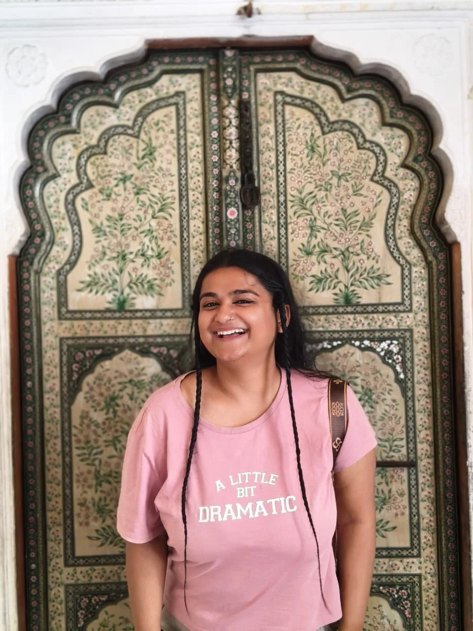A young woman with black hair in a pink T-shirt that says 'A LITTLE BIT DRAMATIC' stands in front of a intricately decorated door with floral and geometric patterns.