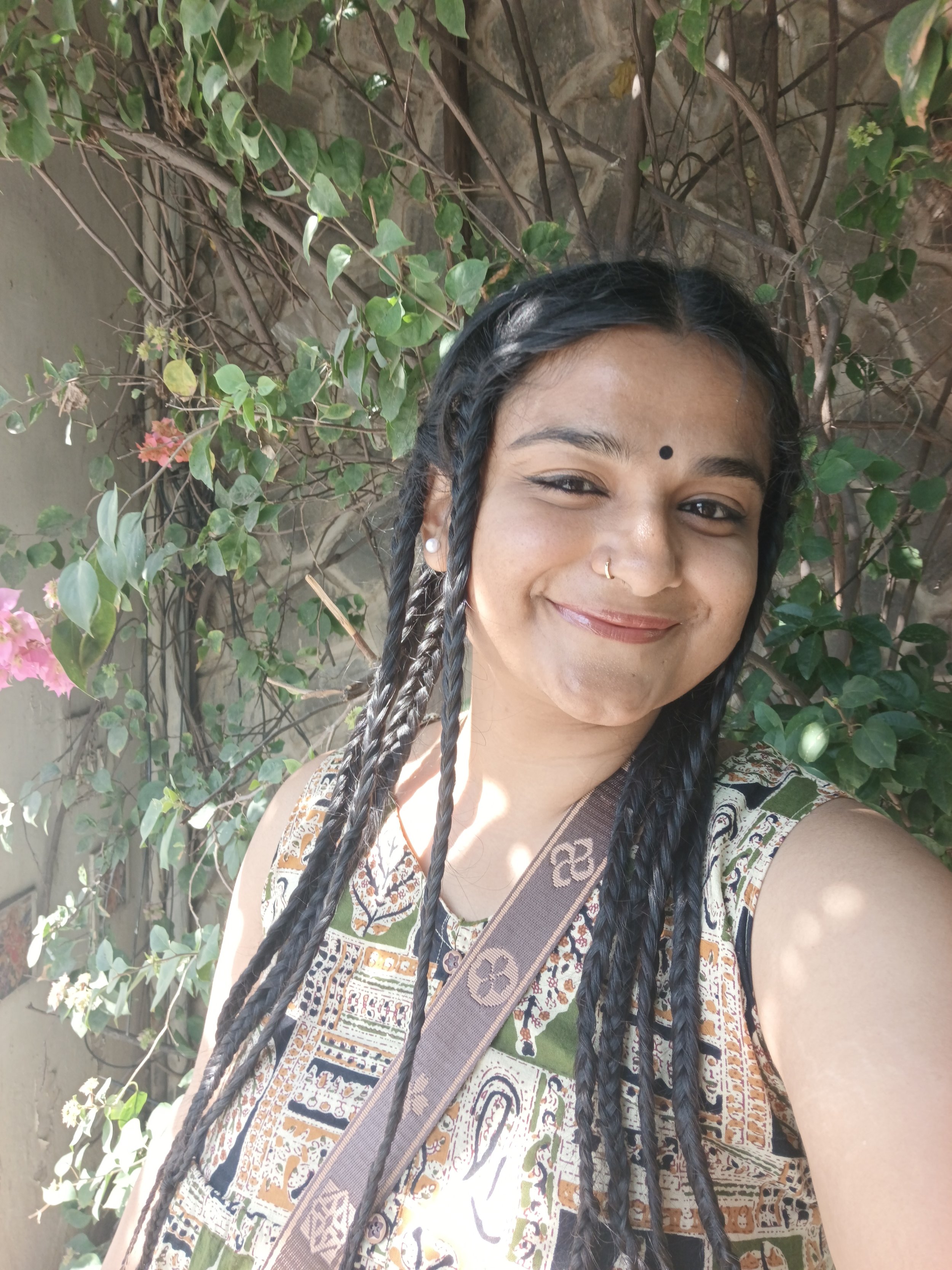 A woman with black braided hair, wearing a patterned sleeveless top, standing in front of a green leafy vine-covered wall, smiling.