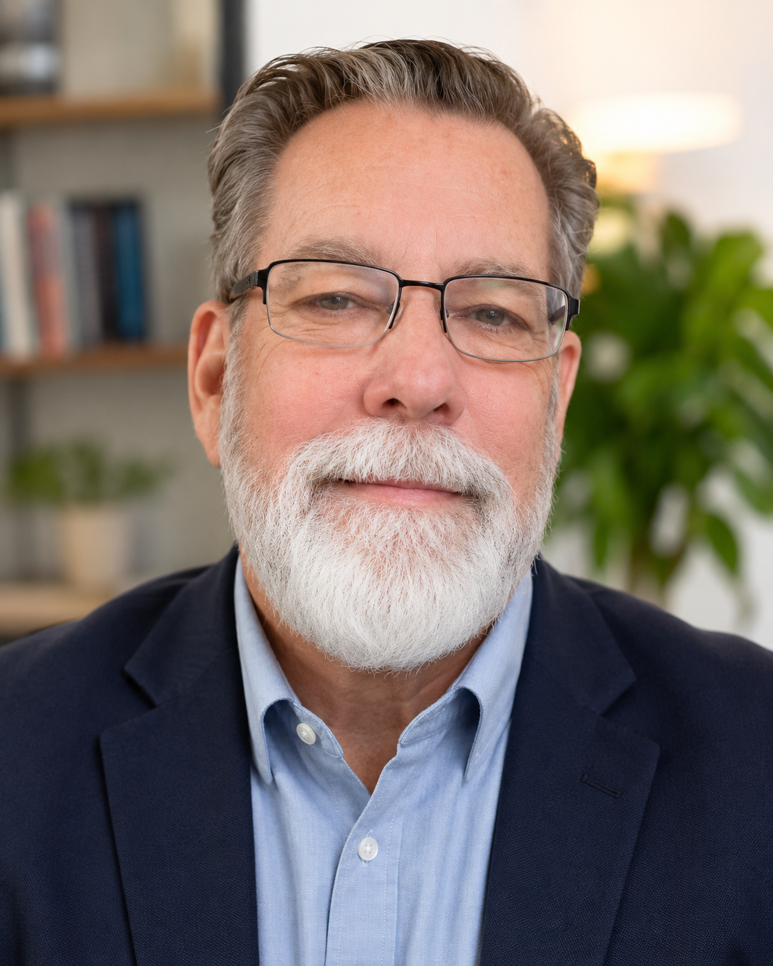 Close-up of an older man with gray hair, beard, and glasses, dressed in a light blue shirt and dark suit jacket, sitting in an indoor setting with blurred bookshelf and a plant in the background.