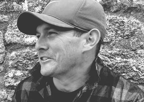 A black and white photo of a smiling man wearing a baseball cap and a plaid shirt, standing in front of a rocky background.