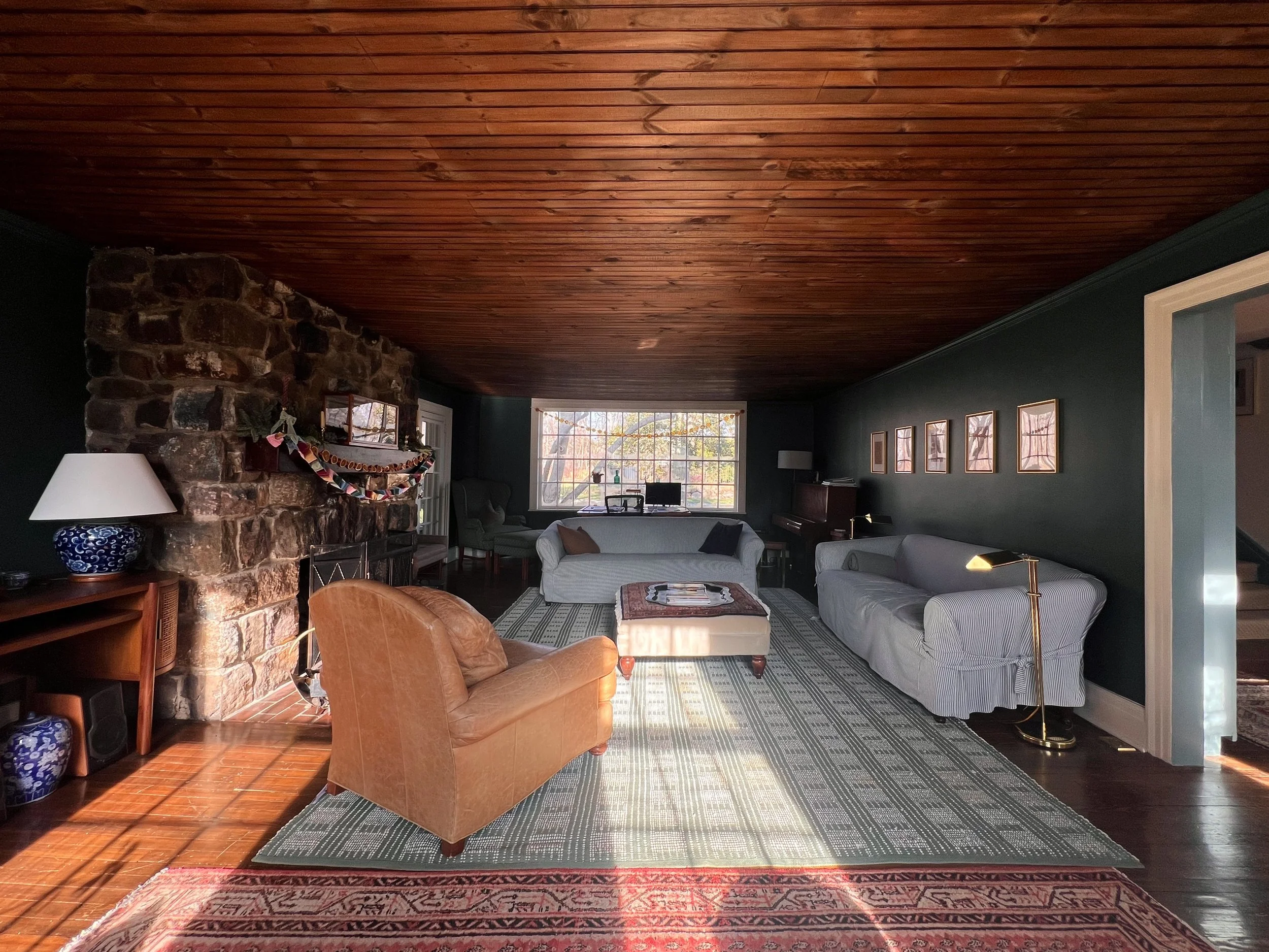 Living room with a wooden ceiling, stone fireplace, dark walls, large window, and various furniture including sofas, a lamp, and framed pictures.