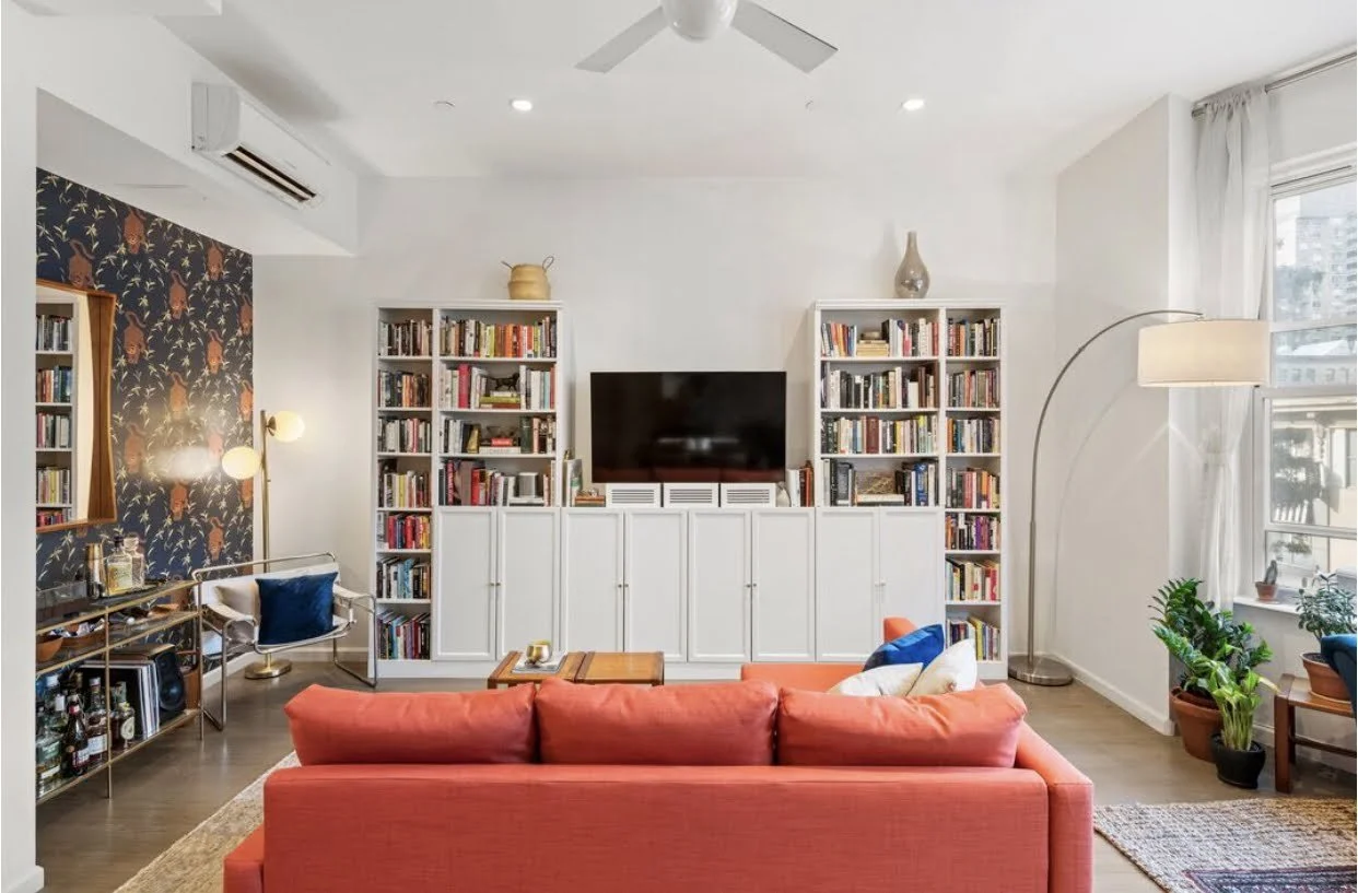 Living room with white walls, a coral sofa, and white bookshelves filled with books. A TV is mounted on the wall between the bookshelves. There is a window with white curtains on the right side, plants by the window, a floor lamp, a small bar cart with bottles and glasses, and a decorative mirror on the left wall.