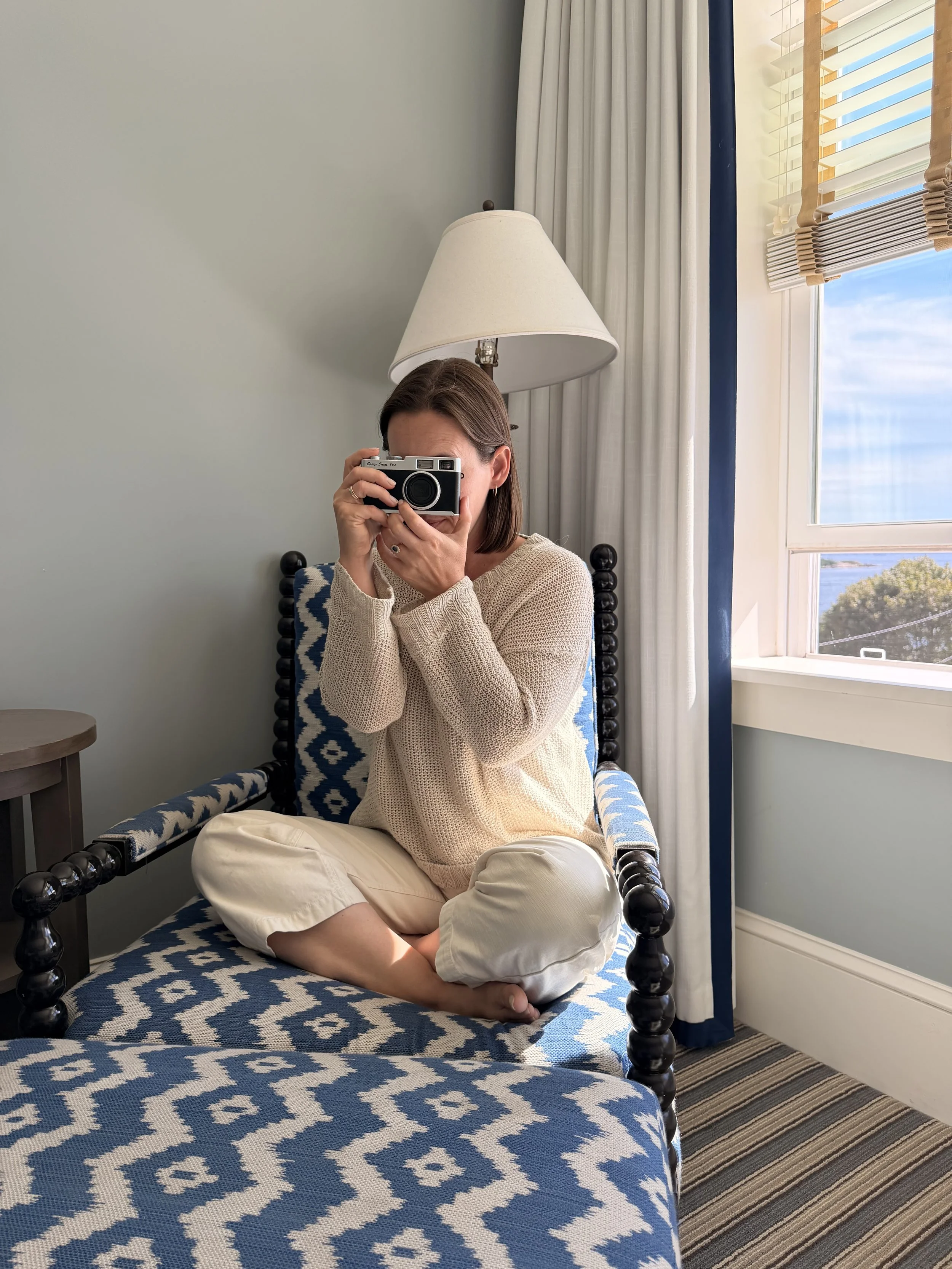A woman sitting cross-legged on a patterned blue and white bed, holding a camera up to her face, in a room with a window, white curtains, and a beige sweater.