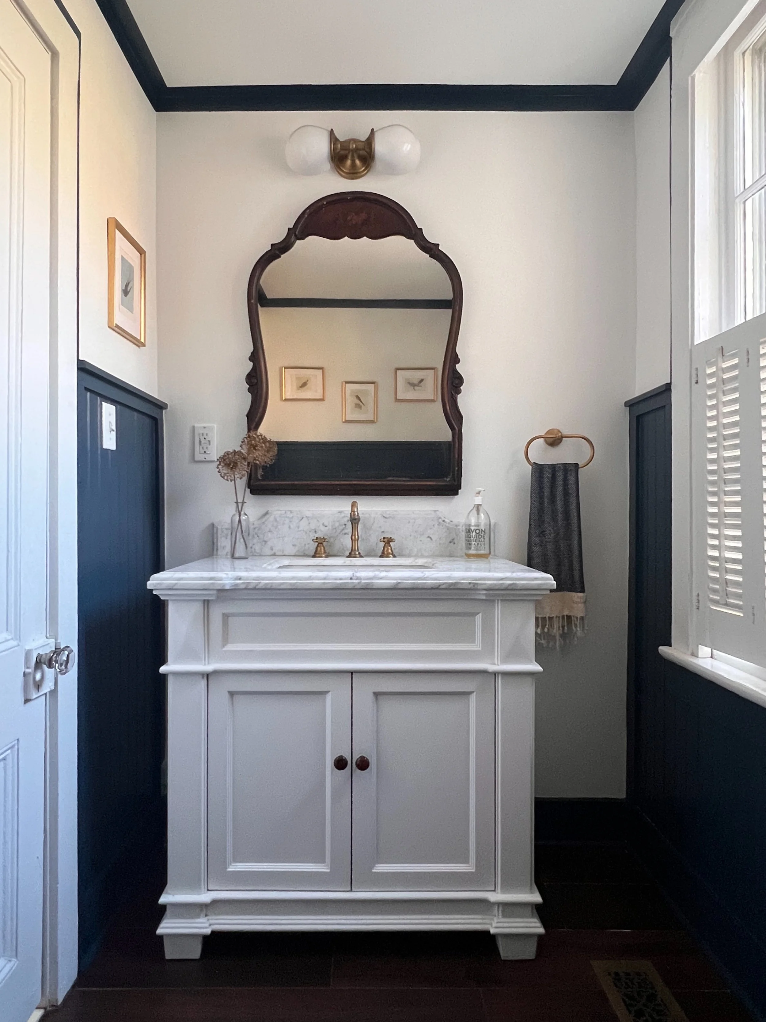 A bathroom vanity with a white cabinet and a marble countertop, a gold faucet, a soap dispenser, and a dried bouquet in a vase. Above the vanity is a large ornamental mirror with a dark wood frame, and three small framed artworks are on the wall behind the mirror. The wall features wainscoting painted in dark color and the upper part painted white, with a brass light fixture above the mirror. To the right, there is a window with shutters.