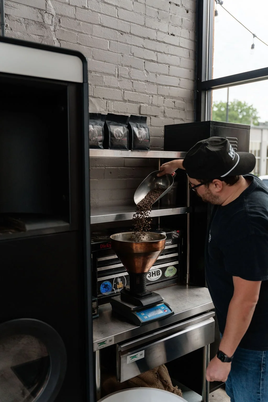 A man wearing a black cap, glasses, and a black t-shirt pours freshly ground coffee beans from a metal scoop into a copper coffee funnel on a digital scale, set on a stainless steel countertop in a modern coffee shop with a brick wall background and a large window.