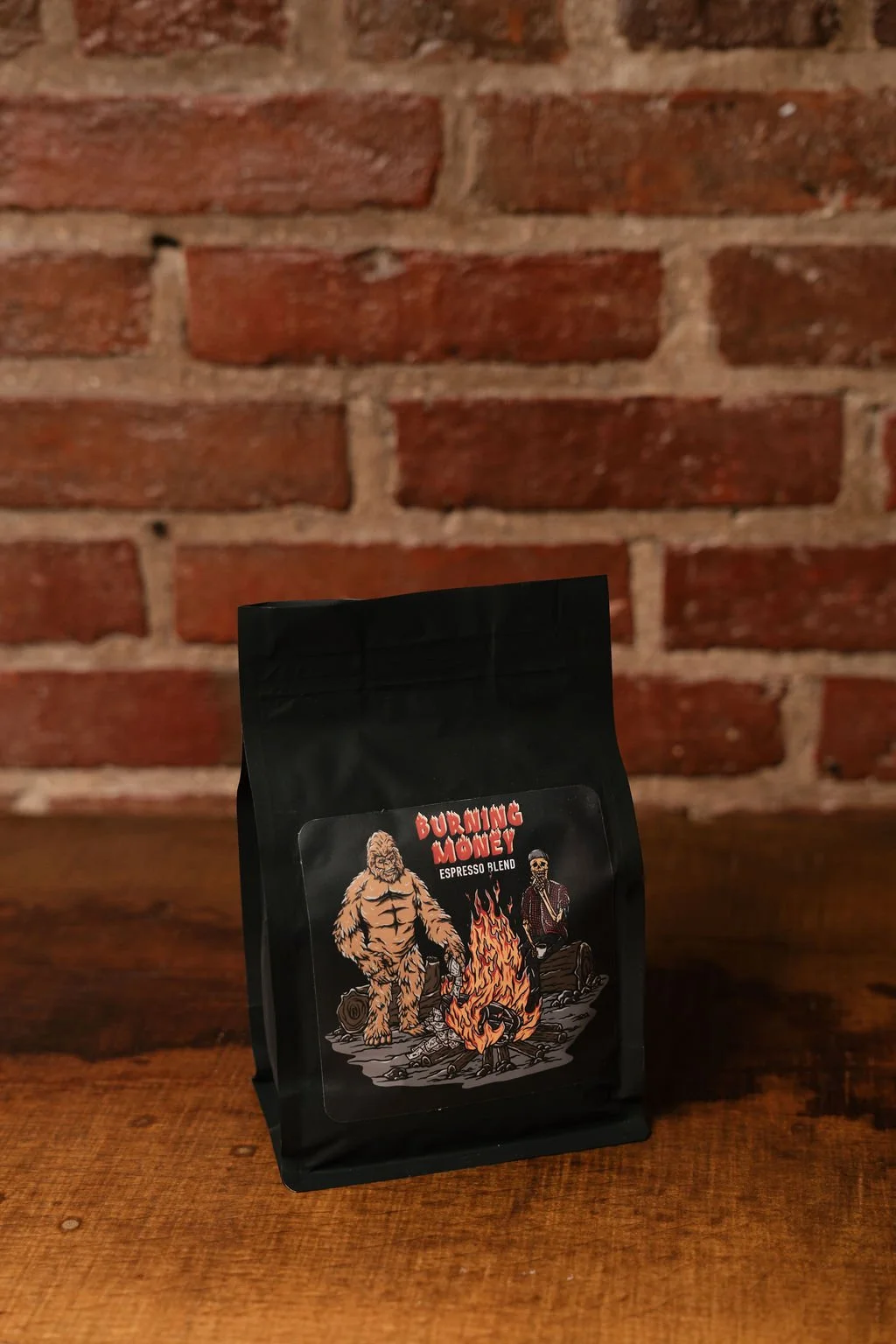 A black bag of coffee labeled Burning Money Espresso Blend with a graphic of a flaming pile of money, a bigfoot figure, and a skeleton sitting next to fire against a brick wall background.