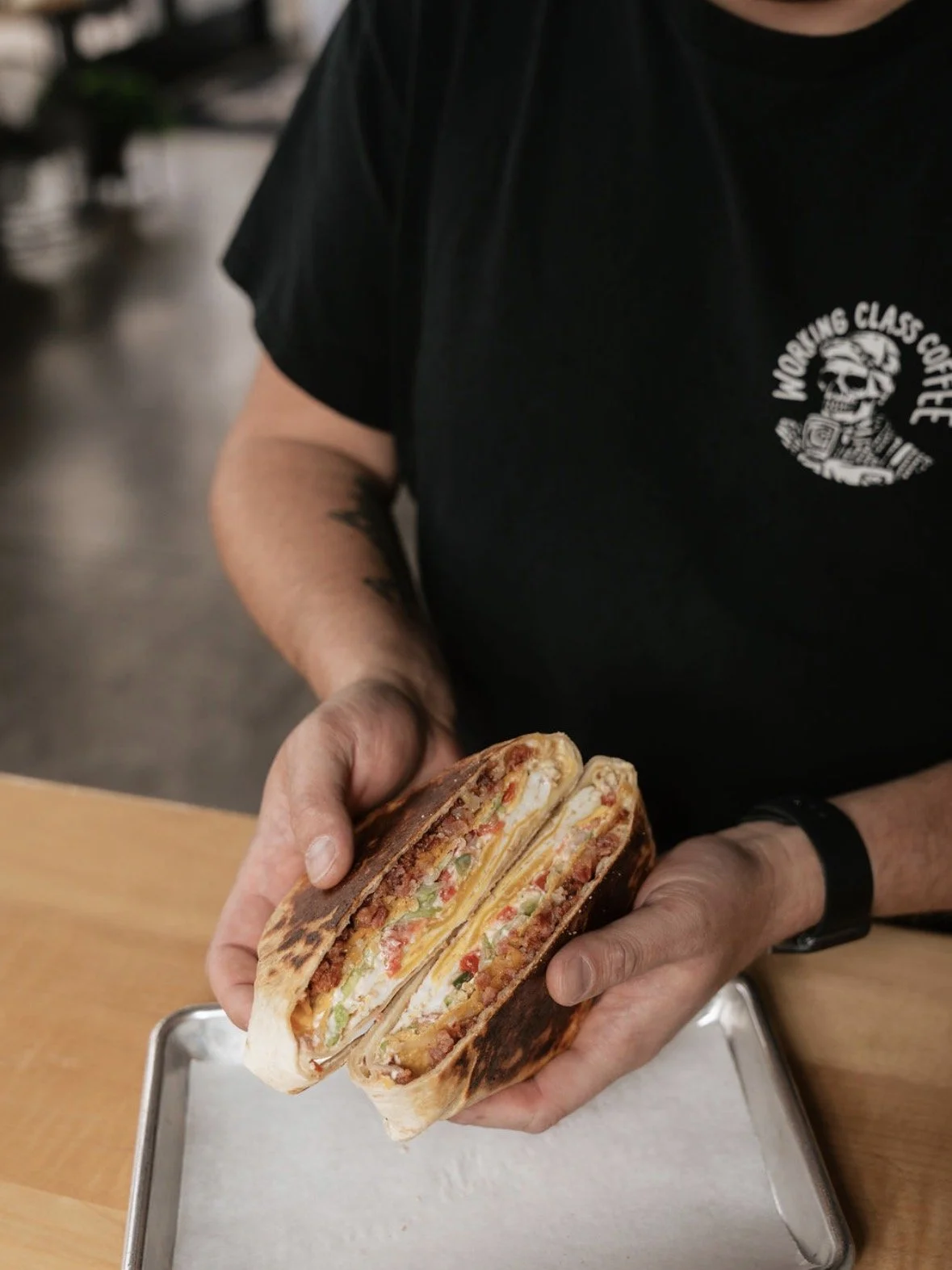 Person holding a half-cut wrap sandwich filled with meat, lettuce, cheese, and vegetables, in a restaurant or cafe setting.