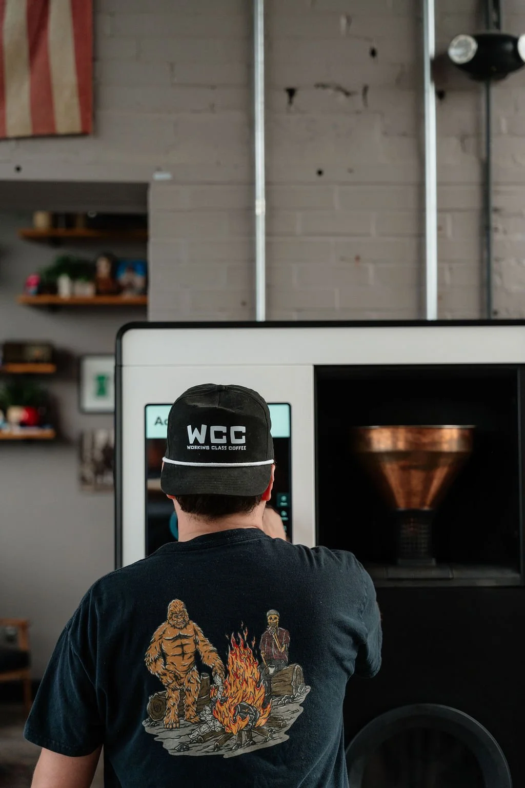 A person from behind wearing a black cap with white text that says 'WCC WORKING CLASS COFFEE' and a black T-shirt with a graphic of a Bigfoot-like creature and a man sitting by a campfire in a coffee shop.