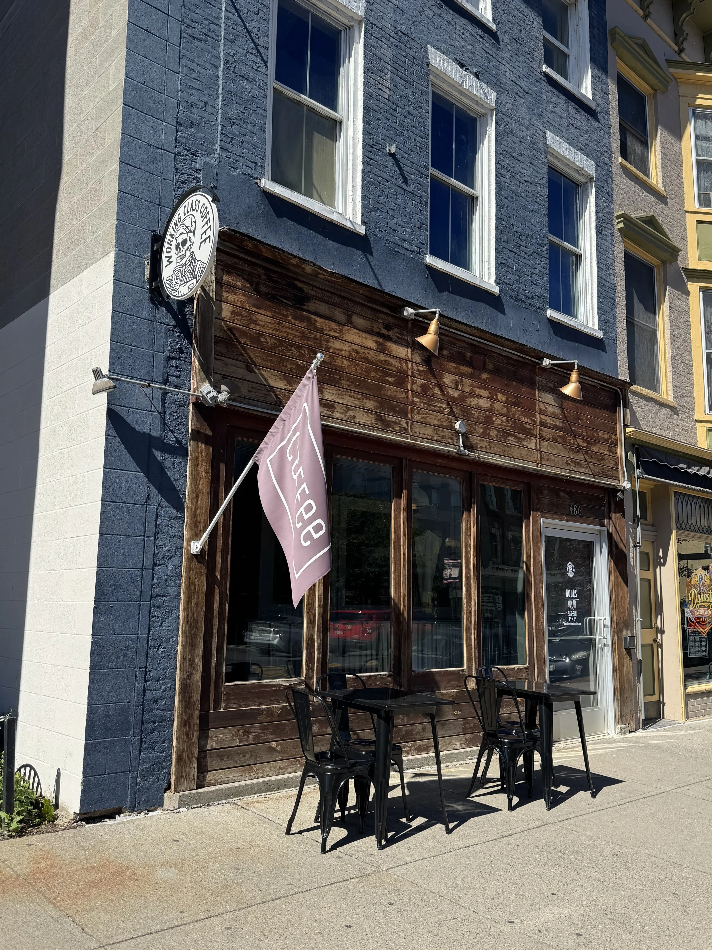 Outside of a coffee shop with black chairs and tables on the sidewalk, a flag with the shop's name, and a sign with a skull and text that reads 'Working Class Coffee' mounted on a building with large windows, dark blue and wooden exterior.