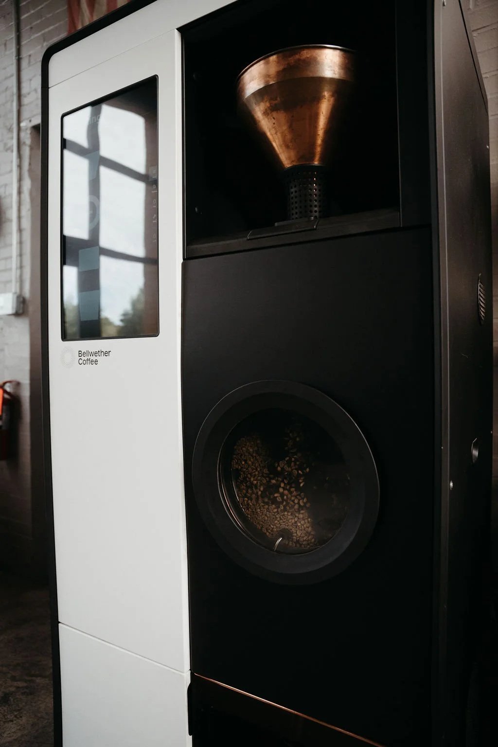 A coffee roaster with a glass door showing coffee beans inside, a large cone-shaped grinder on top, and the label 'Bellwether Coffee' on the front.