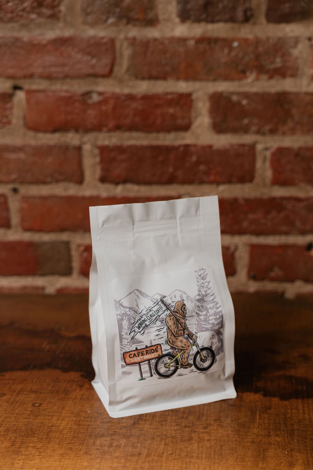 White coffee bag with a mountain and forest illustration, featuring a Sasquatch riding a bicycle, in front of a brick wall background.