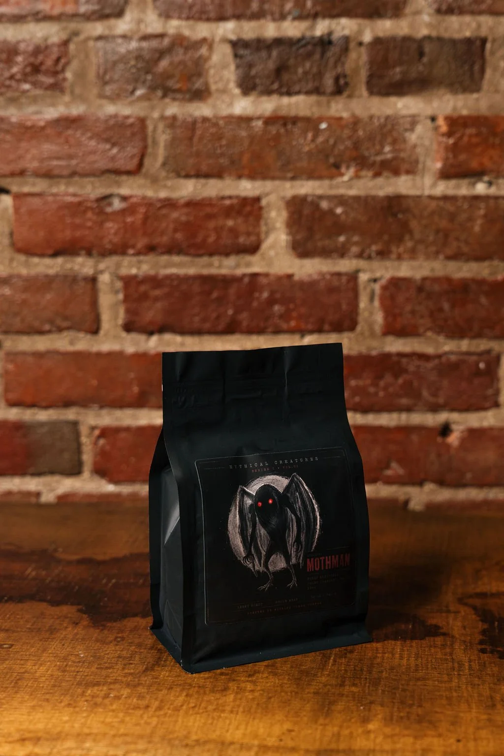A black bag of Mothman coffee with a graphic of a creature with red eyes and wings, placed on a wooden surface against a brick wall background.