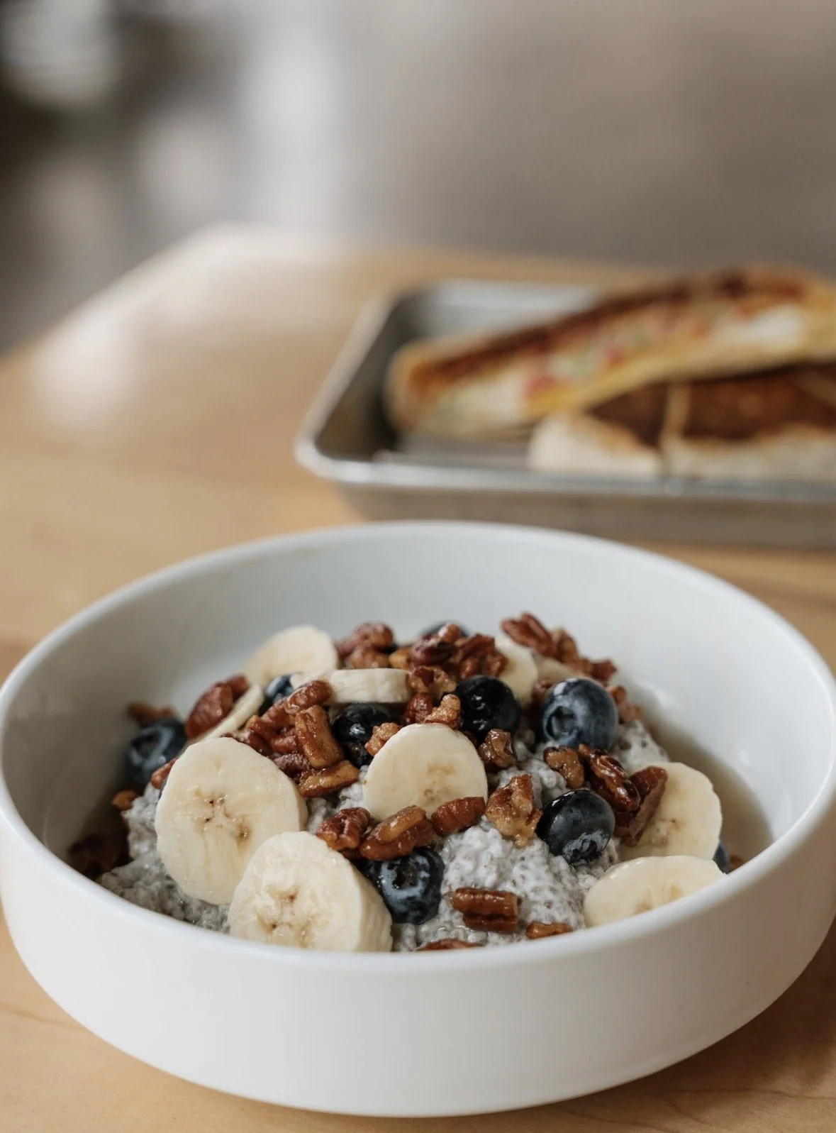 Bowl of chia pudding topped with banana slices, blueberries, and chopped pecans, with a plate of grilled sandwiches in the background.