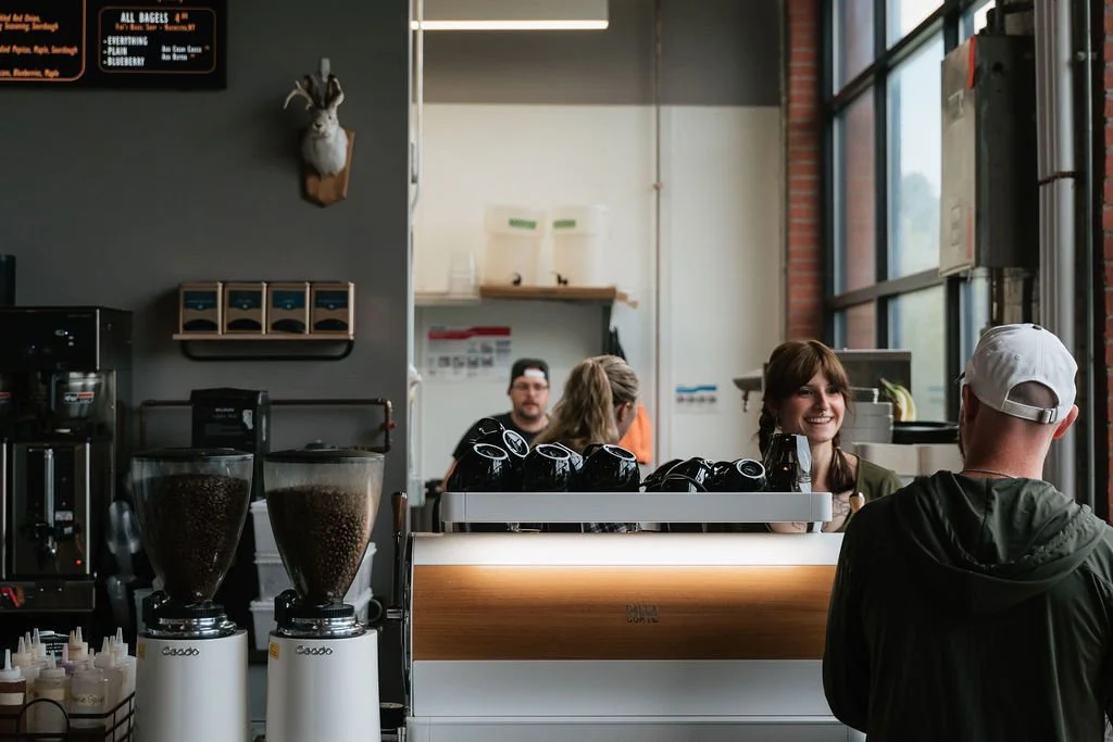 Coffee shop counter with two coffee grinders, a customer wearing a white cap and black jacket, and two baristas behind the counter smiling. Large windows let in natural light.