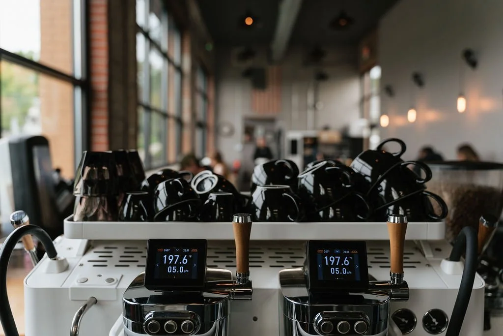 Commercial espresso machine with dual group heads, displaying temperature and pressure, filled with black coffee mugs, inside a coffee shop with large windows and blurred customers in background.