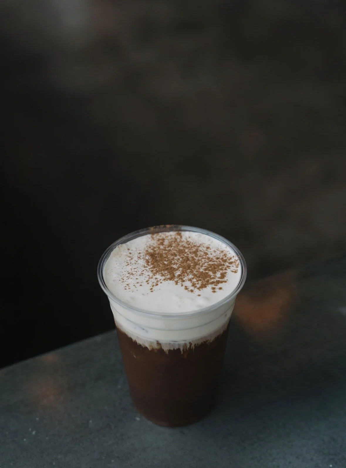A tall glass of iced coffee with whipped cream and cocoa powder on top, placed on a dark surface.
