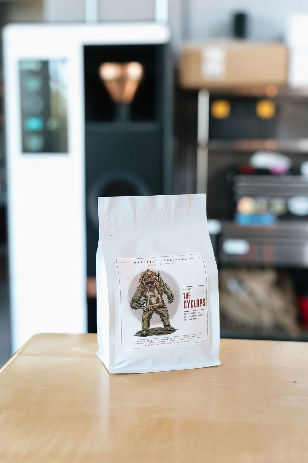 A white package labeled 'The Cyclops' on a wooden table with a cartoon monster illustration on the label. Blurred kitchen or storage area in the background.