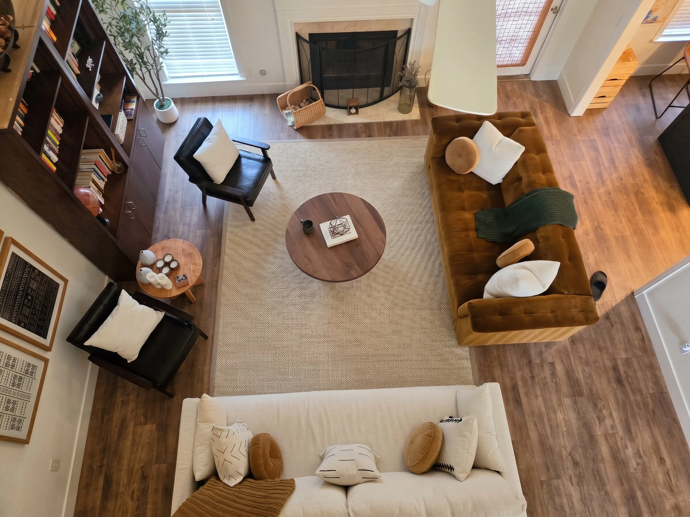 Living room with a brown velvet sofa, white chair, black chair, round wooden coffee table with decor, fireplace, large window with blinds, bookshelf, and hardwood floors.