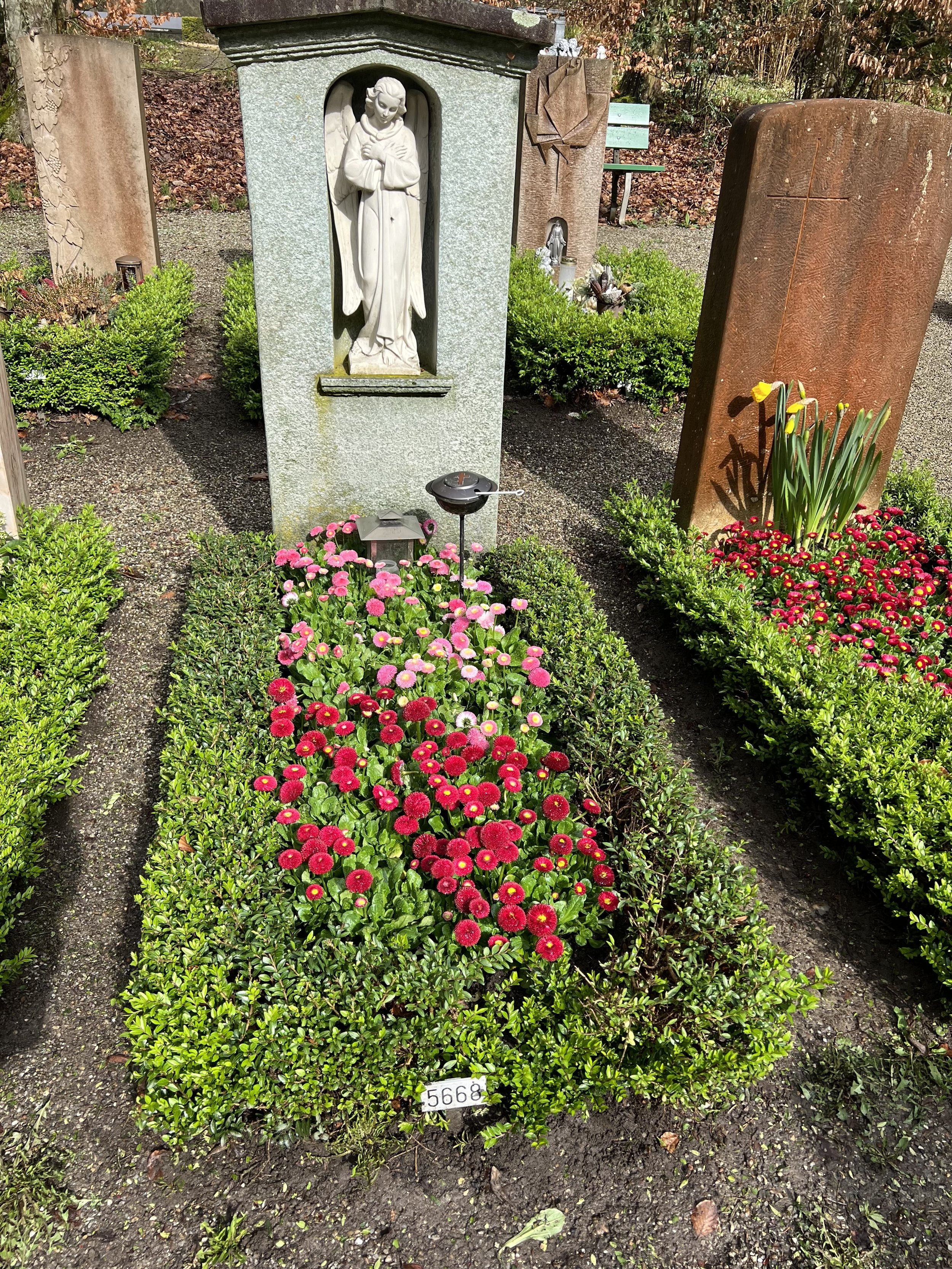 Grabstätte mit einer geschnitzten Marienfigur in einer Grabstätte, umrahmt von grünen Blumen und bunten Chrysanthemen, auf einem Friedhof im Herbst.