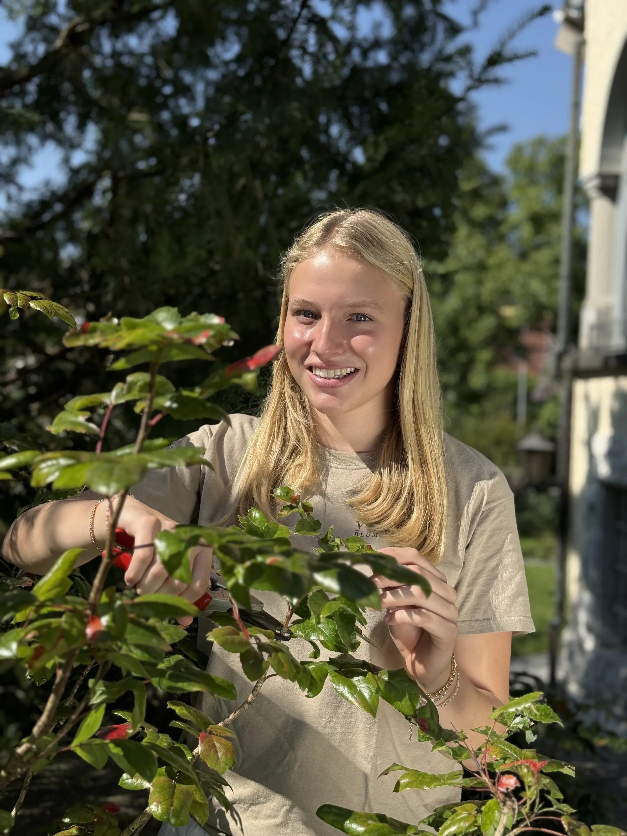 Eine junge blonde Frau pflückt rote Beeren an einem Strauch im Garten bei Sonnenlicht.