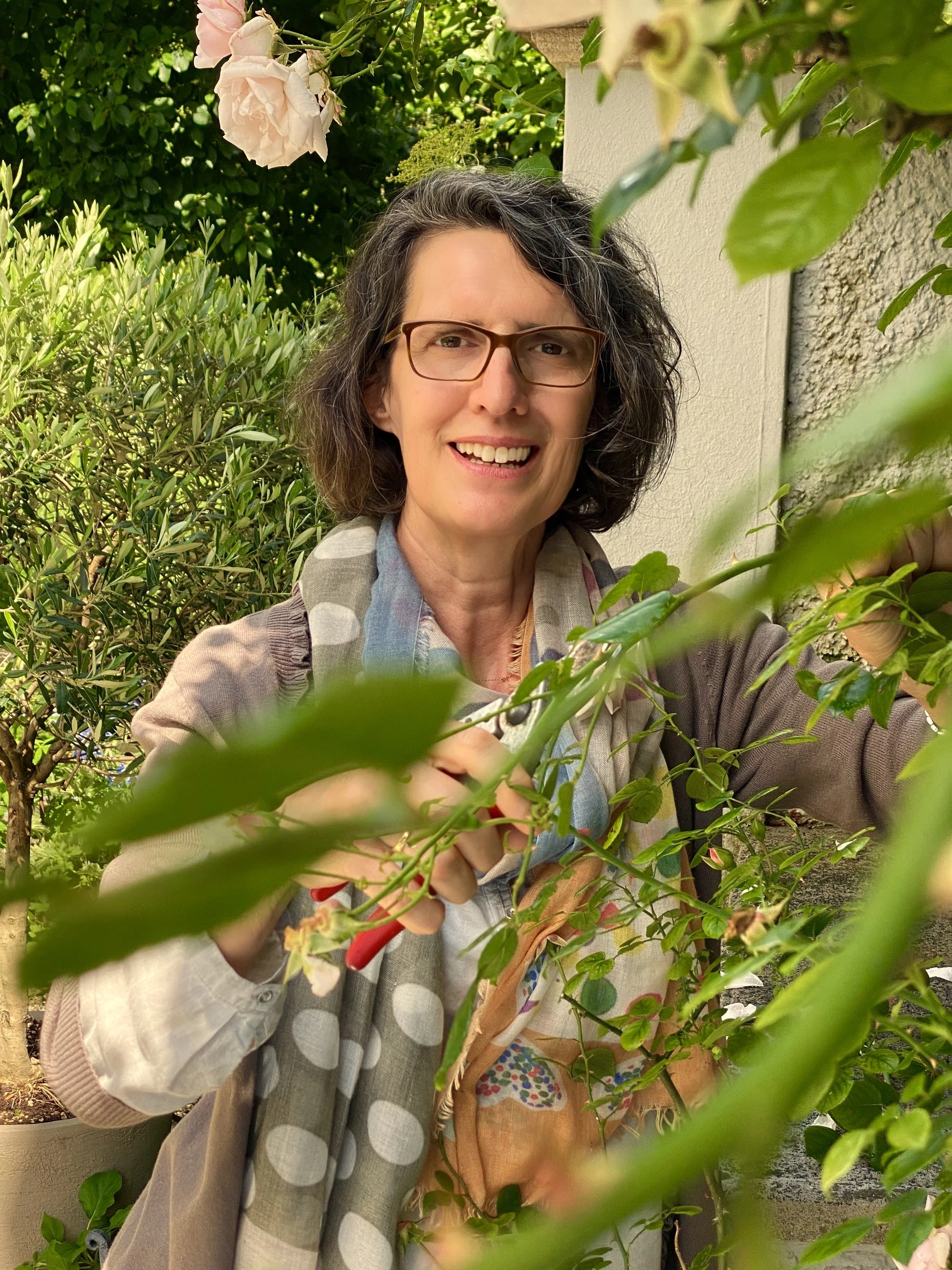 Eine Frau mit Brille beim Schneiden einer Pflanze im Garten, umgeben von grünen Blättern und einer weißen Wand im Hintergrund.