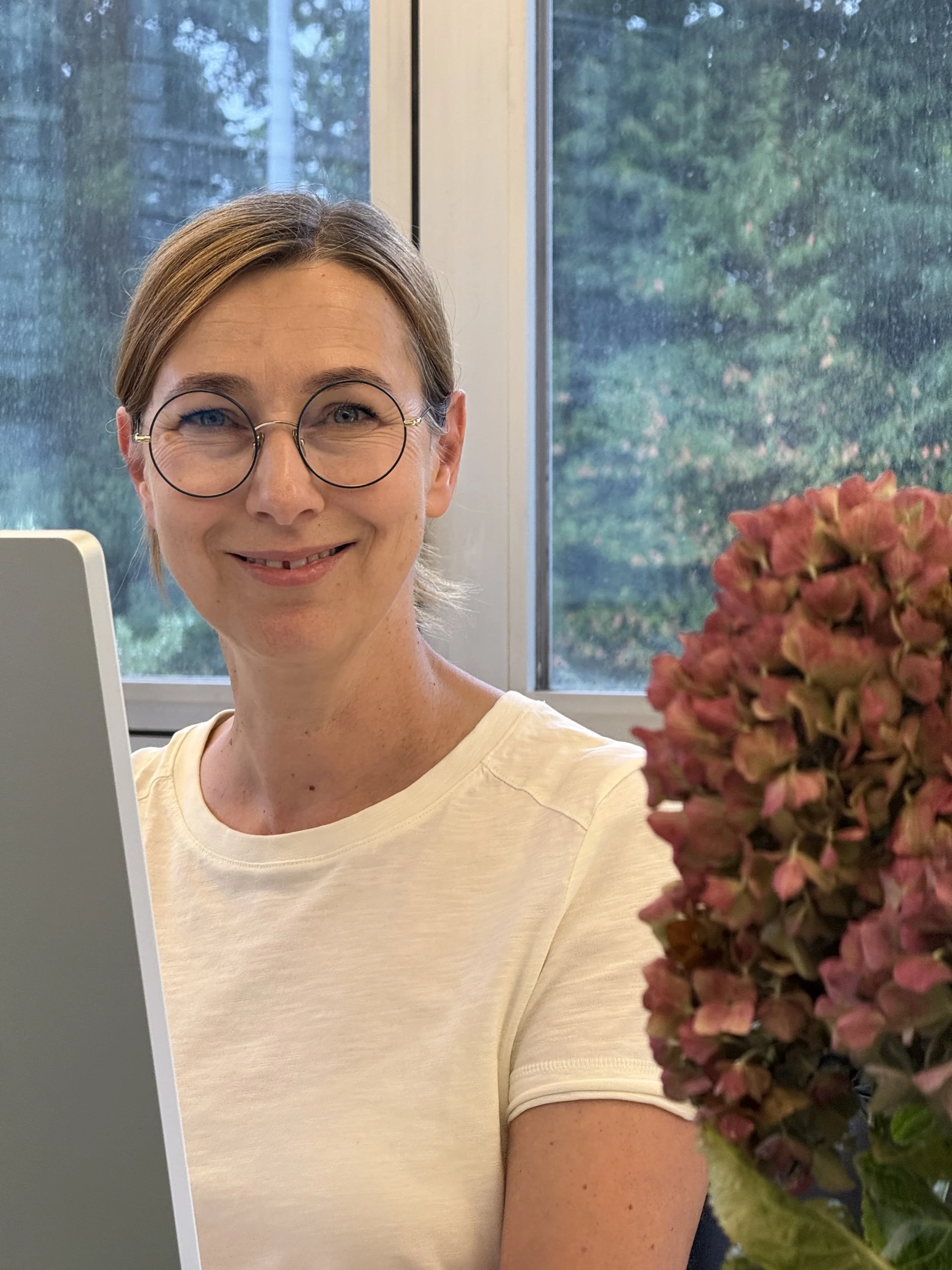 Ein freundliches Frau trägt eine weiße T-Shirt, trägt runde Brille und hat kurze, braune Haare. Sie sitzt vor einem Fenster mit Blick auf Bäume. Neben ihr befindet sich eine Topfpflanze mit rosa Blüten.