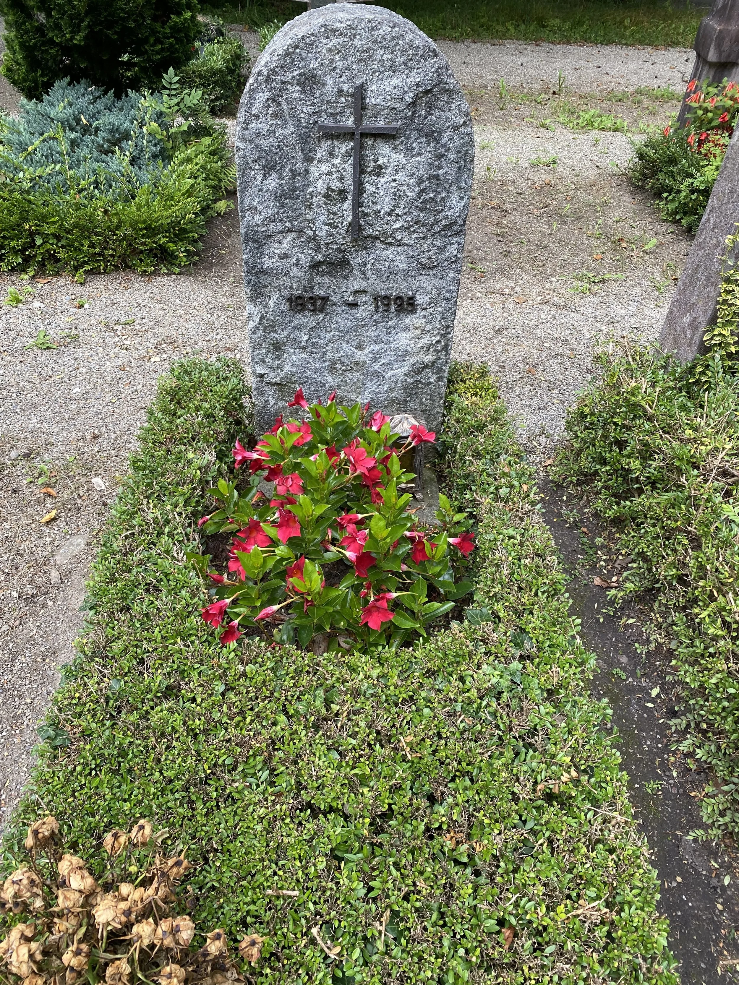 Ein Grabstein aus grauem Stein mit einer schwarzen Metallkreuz-Statue auf der Oberseite, beschriftet mit den Jahren 1997 – 1995, umgeben von bunten Blumen und grünen Sträuchern.
