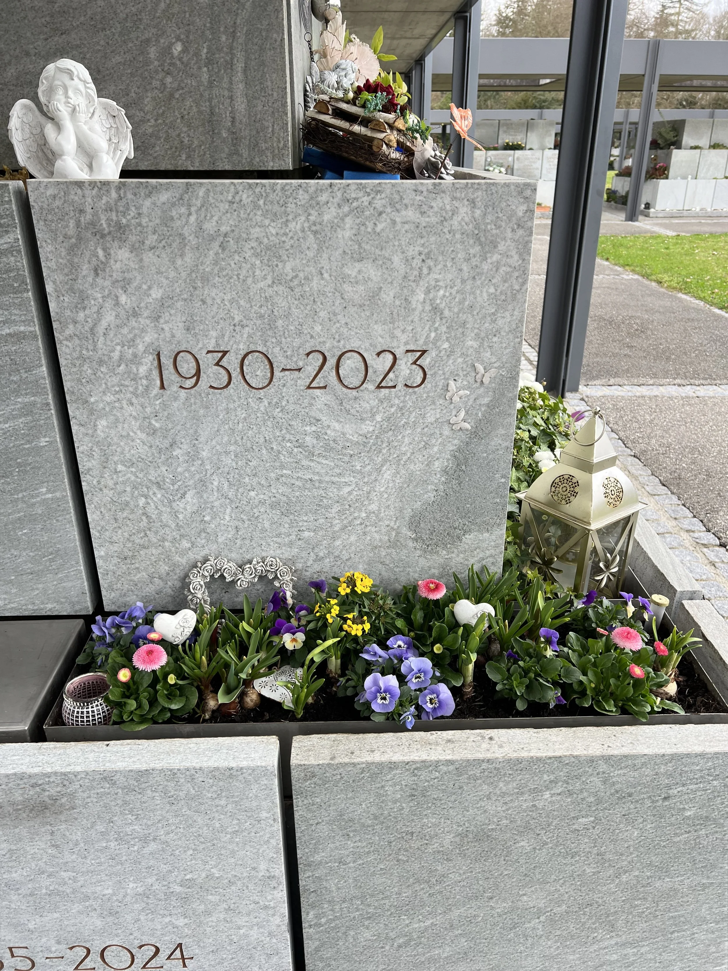 Grabstein mit den Jahren 1930-2023, umgeben von bunten Blumen, Kerzen und einer kleinen Laterne, auf einem Friedhof.